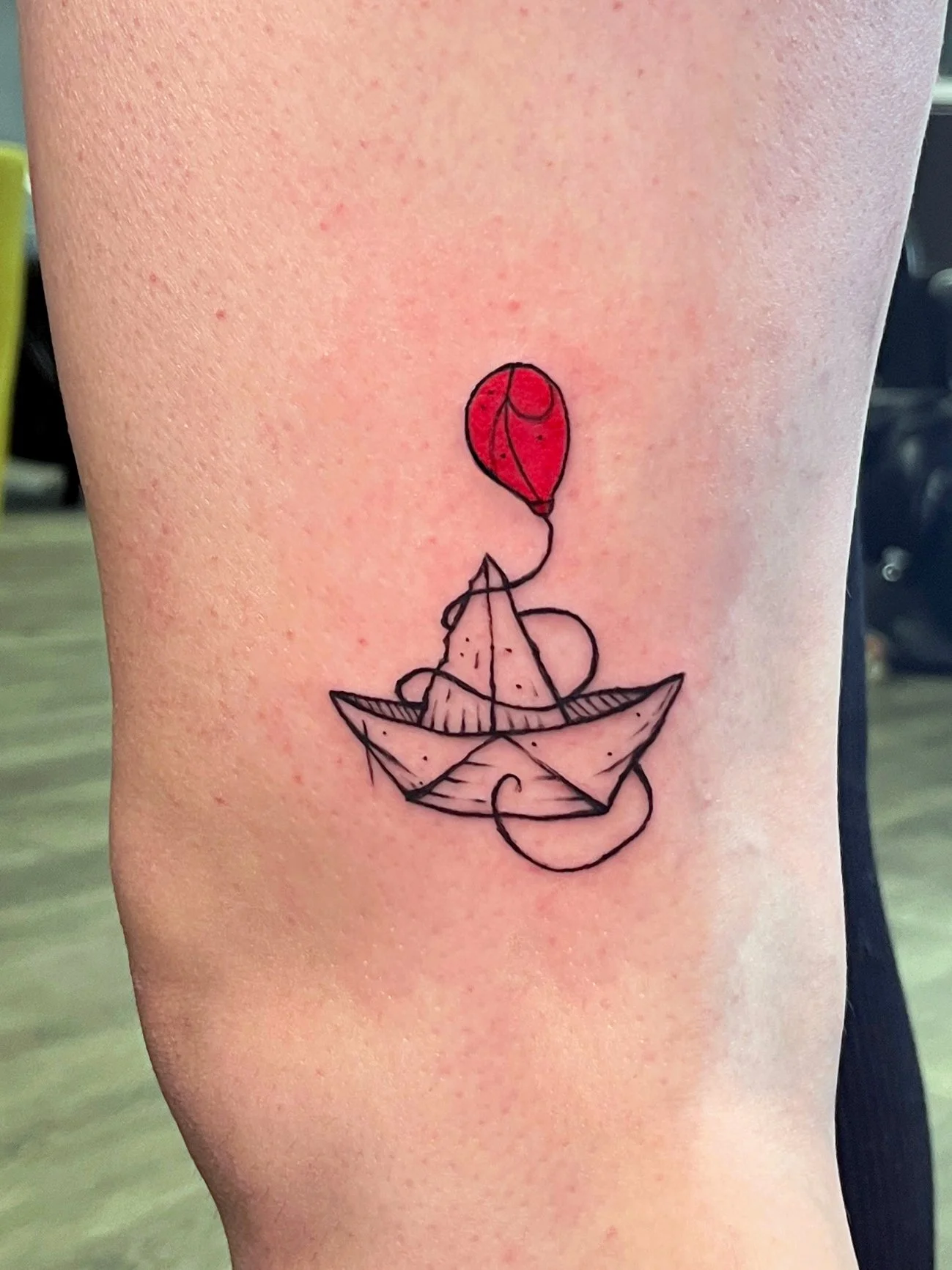 A tattoo of a simple line drawing of a paper boat with a red balloon tied to the top of the boat's mast.