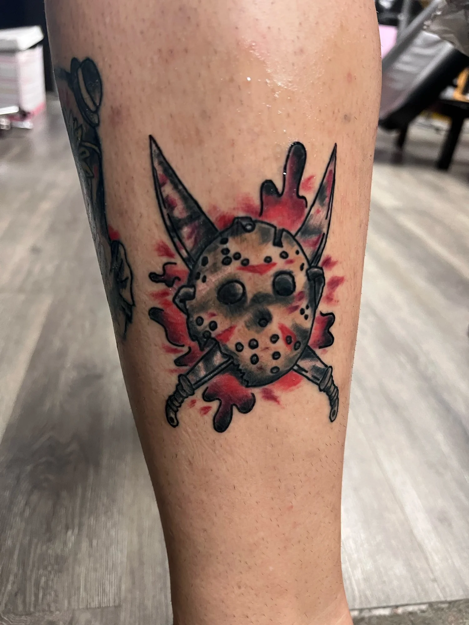 Tattoo of a hockey mask with mechanical limbs and two knives crossed behind it, with red paint splatters in the background.