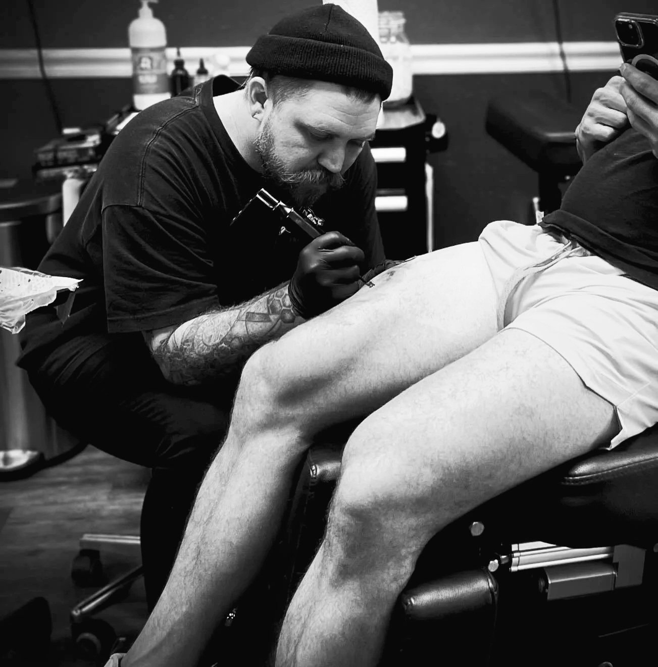 Desmond Carew in a beanie and gloves is tattooing the thigh of a person lying face down on a chair in a tattoo studio.