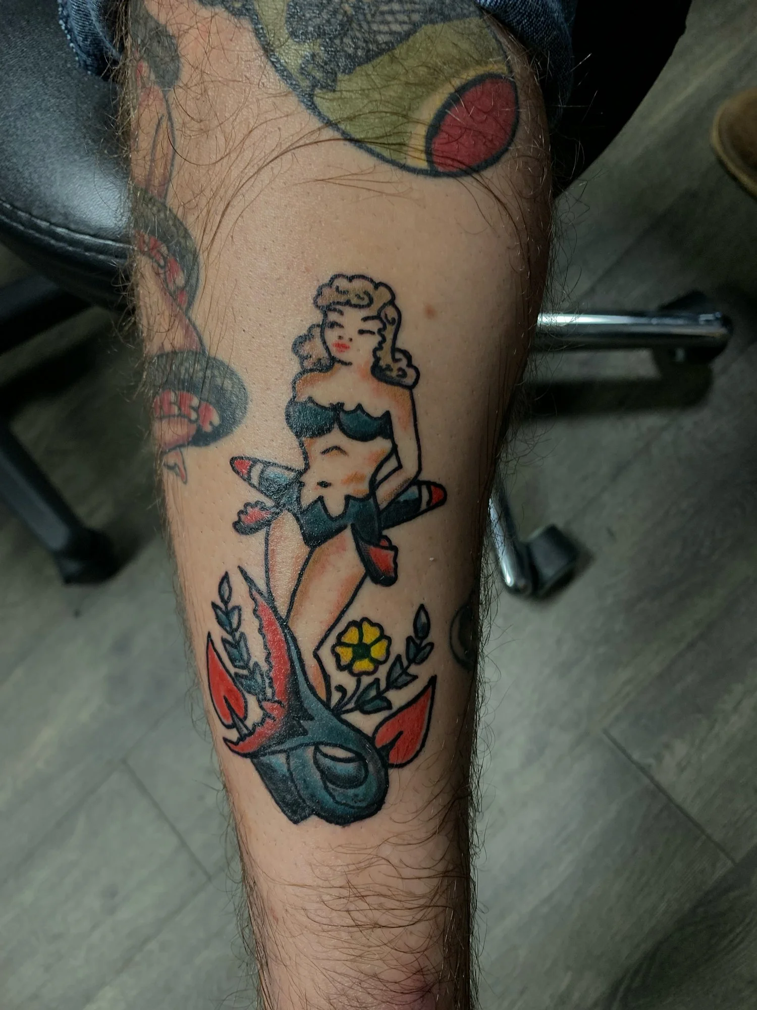 Tattoo of a vintage-style woman with blonde hair and red lipstick, sitting on a large fish or sea creature, with small flowers at the bottom of the tattoo.