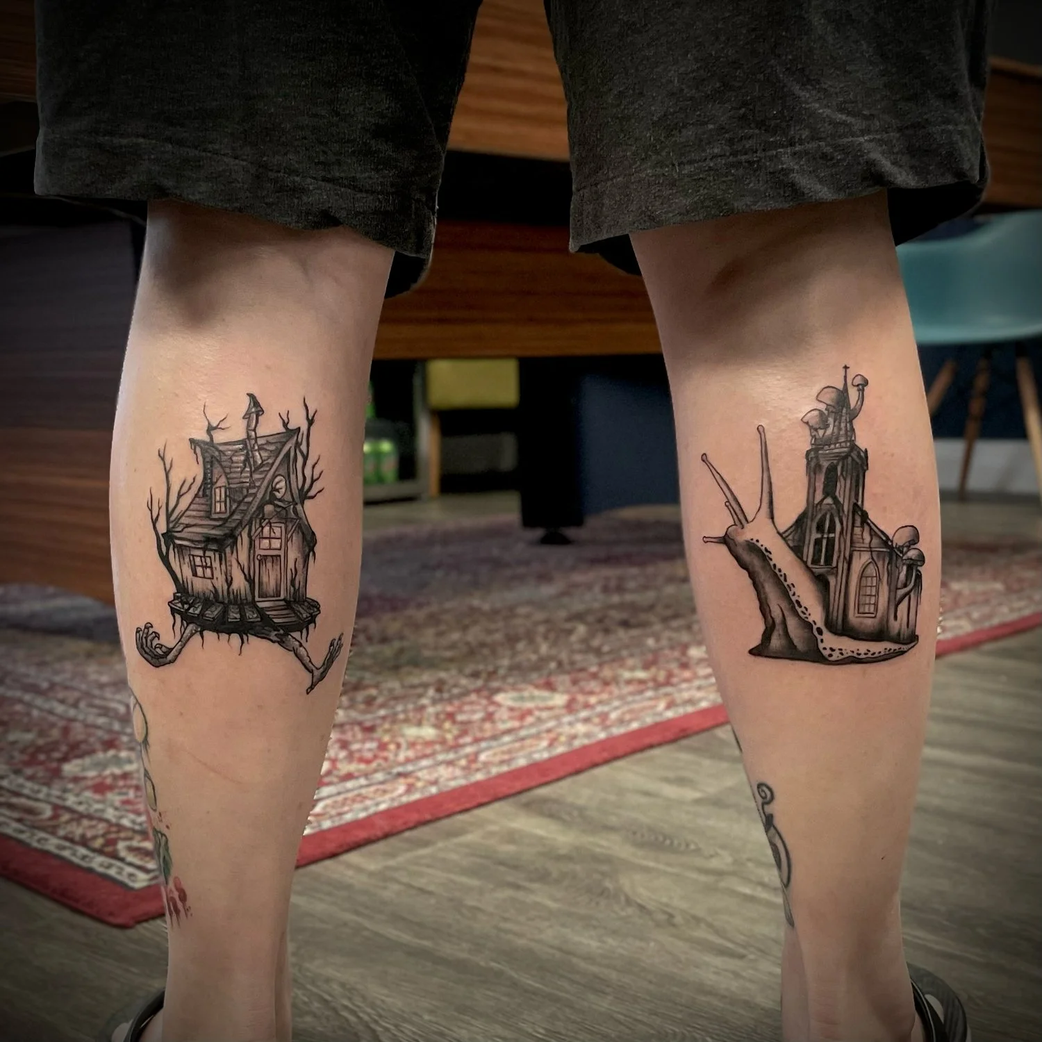 Photo of two legs with house-themed tattoos on the calves, featuring haunted houses with trees and spooky elements, in an indoor setting.