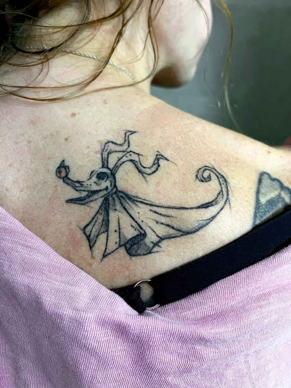 A tattoo of a whimsical dragon with wings on a person's upper back near the shoulder, with wet hair and a necklace visible.