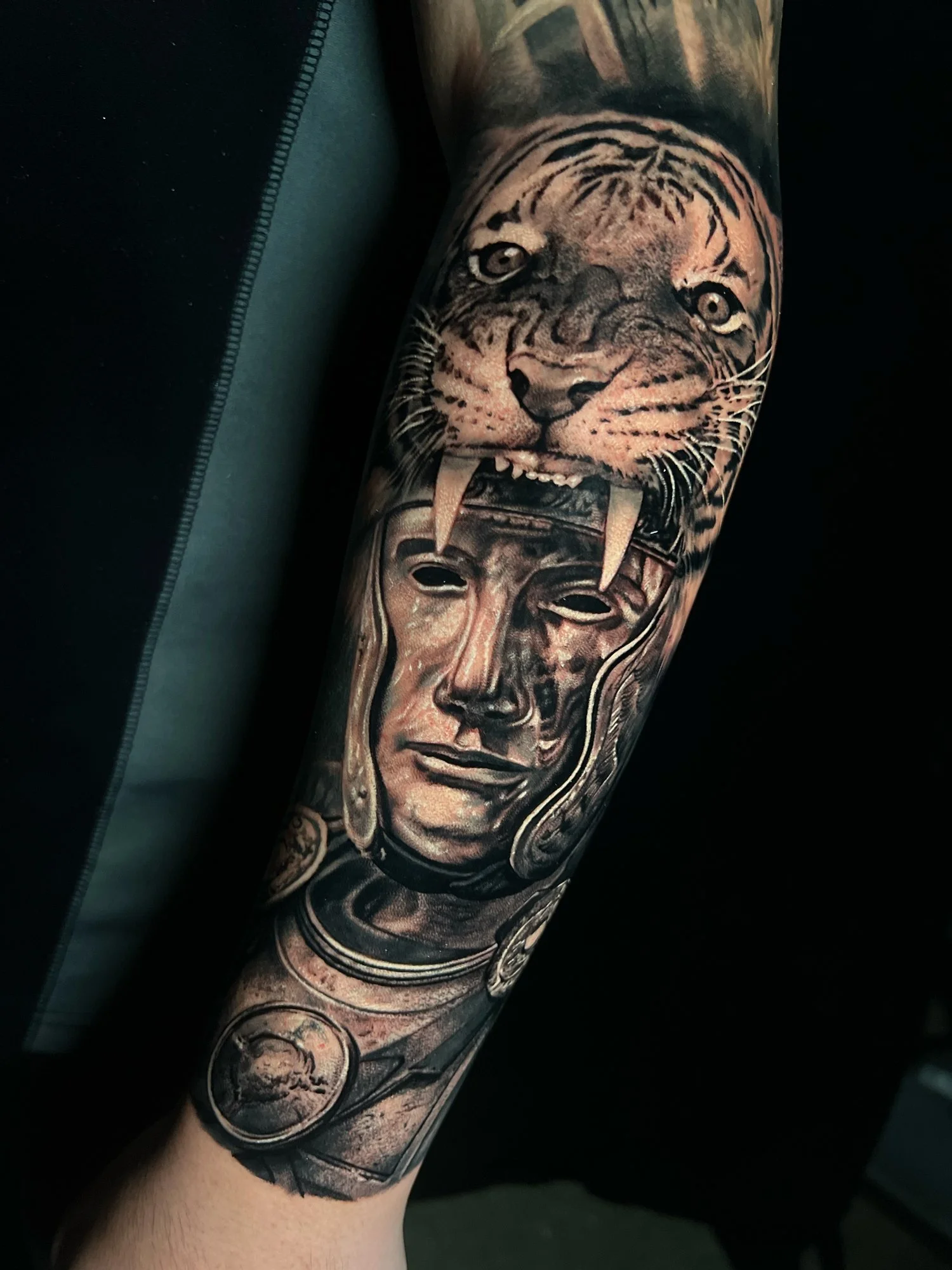 Black and grey tattoo of a fierce tiger with sharp teeth above a realistic Greek goddess head sculpture with flowing hair on a forearm.