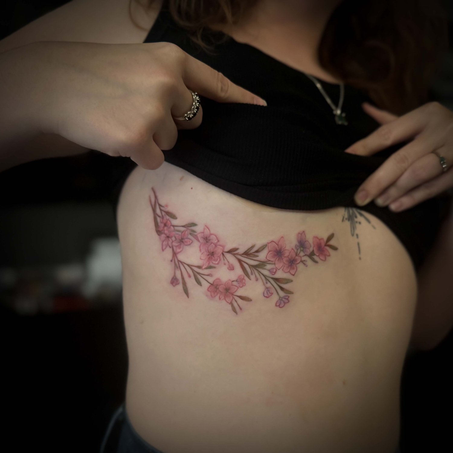 A person lifting their black top to reveal a fresh, colorful floral tattoo on their upper thigh.