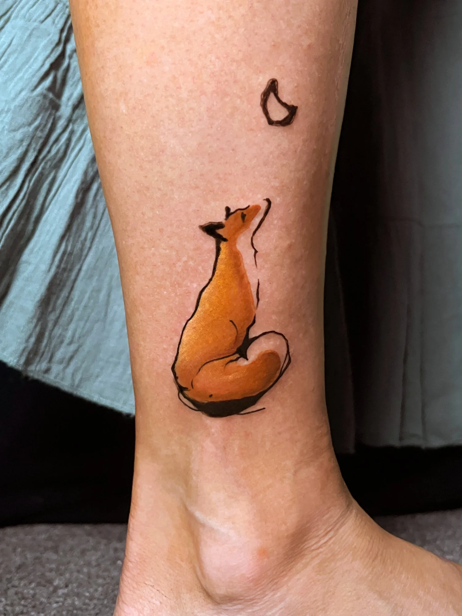 Tattoo of a sitting fox with a moon above its head on a person's leg.