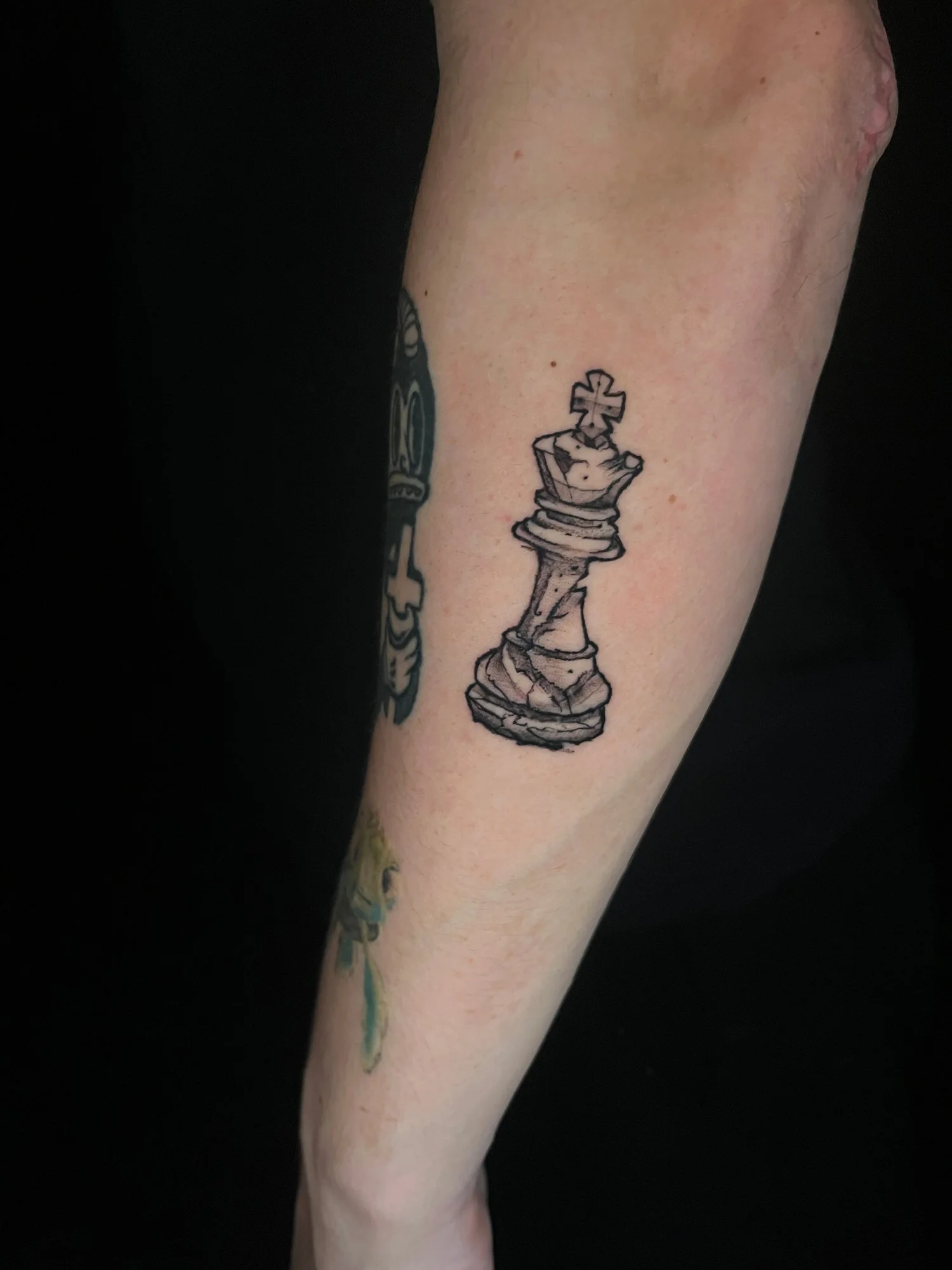 Black and gray tattoo of a chess king piece on the forearm.