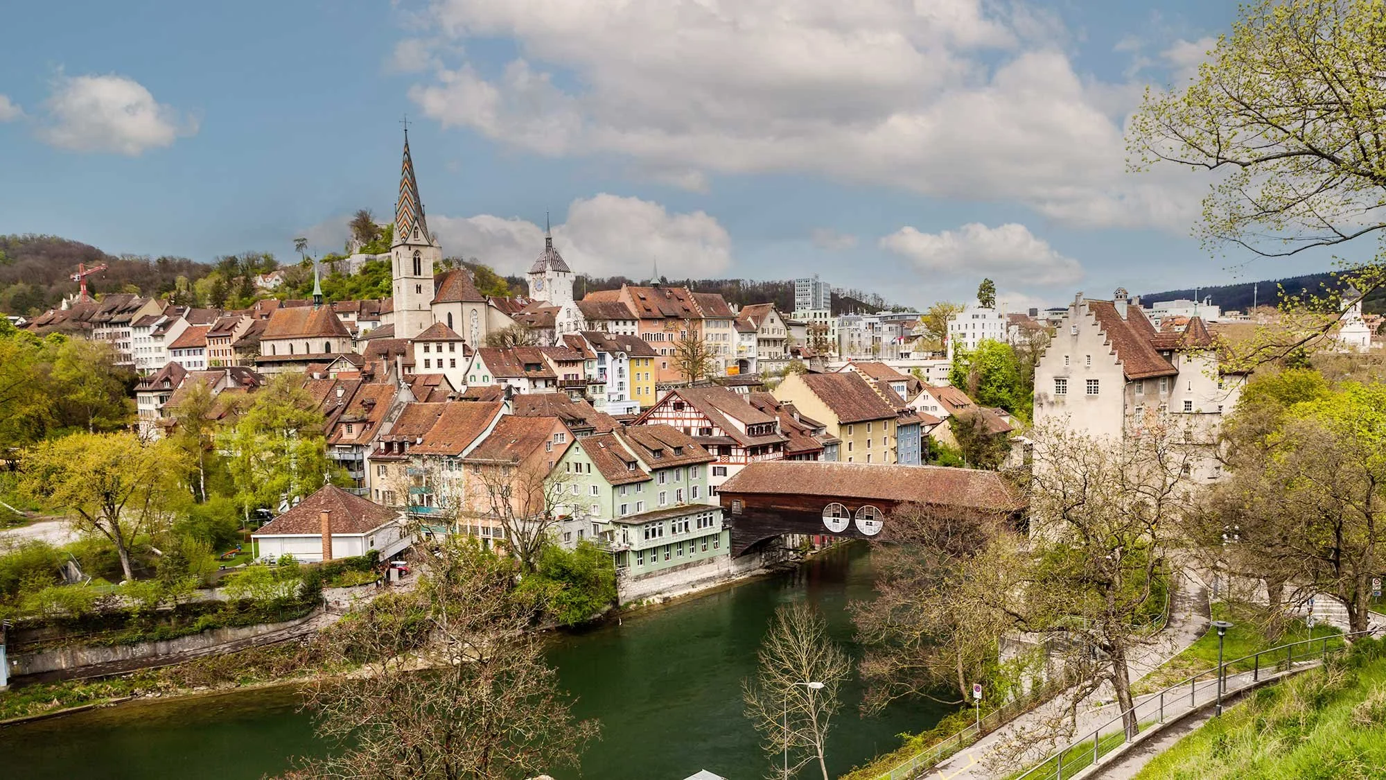 Ein Stadtbild von Baden, im Kanton Aargau mit historischen Gebäuden und Kirchen, die Limmat im Vordergrund, grüne Bäume und bewölkter Himmel.