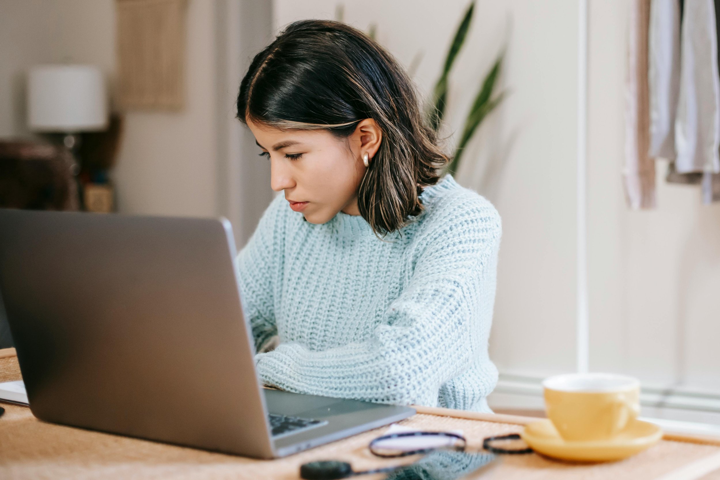Woman sitting at a table working on a laptop with a cup and hair clips on the table.
