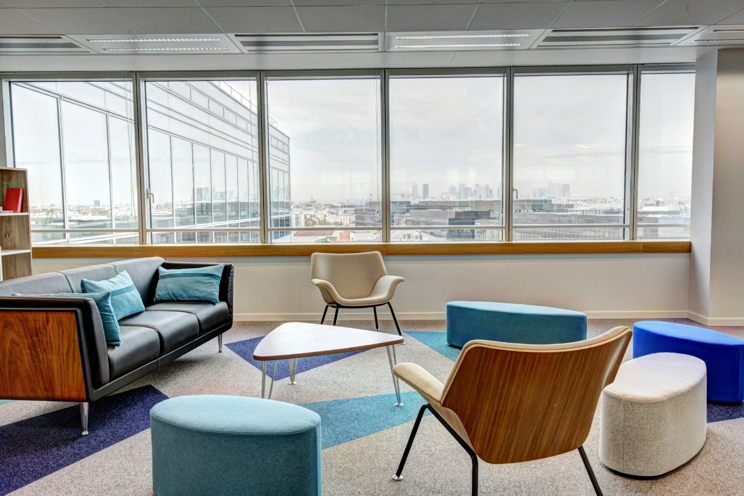Modern office lounge with colorful ottomans, chairs, and a couch, large windows showcasing a city skyline.
