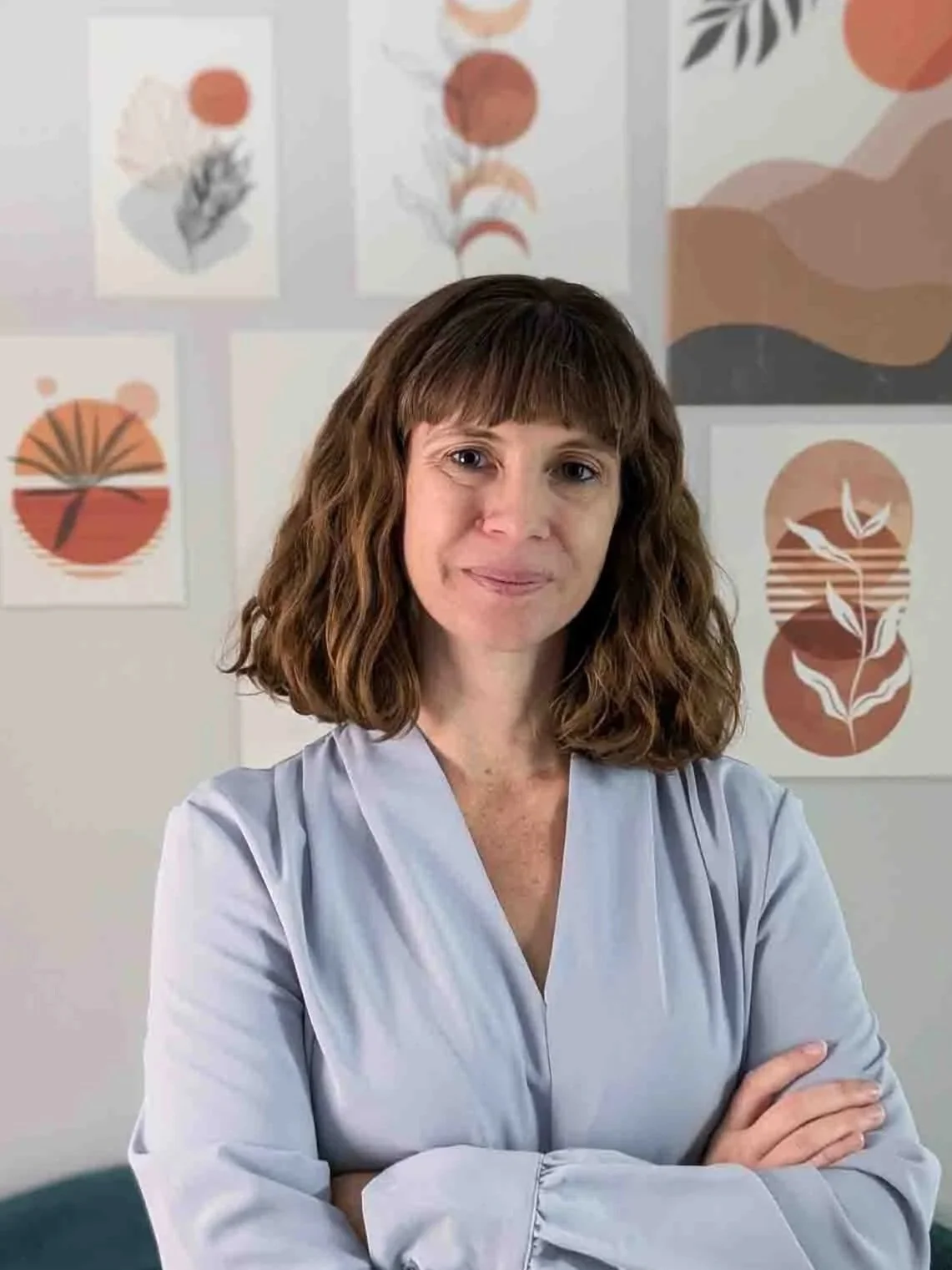 A woman with wavy brown hair and wearing a light gray blouse, standing with arms crossed in front of a wall decorated with abstract art in shades of beige, orange, and gray.
