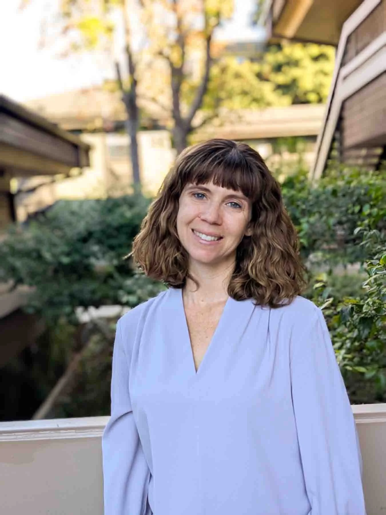 A woman with shoulder-length curly brown hair and bangs, wearing a light blue blouse, smiling outdoors with houses, trees, and bushes in the background during daytime.