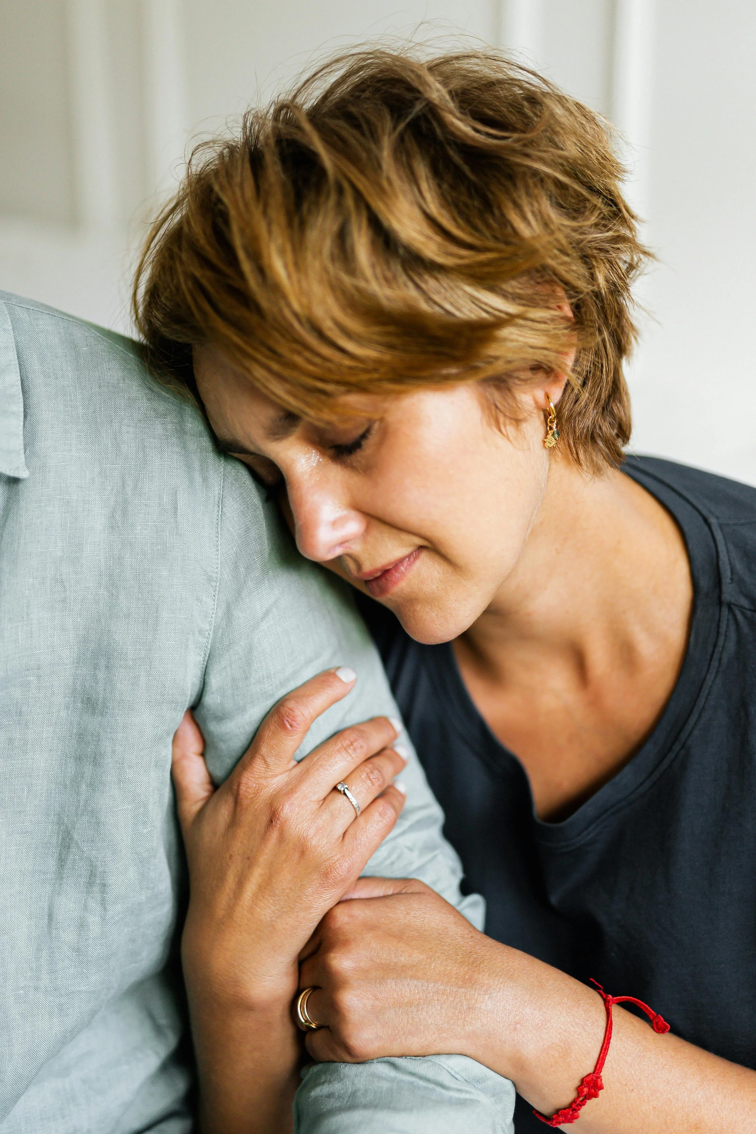 A woman with short reddish hair and earrings is resting her head and hand against a person's shoulder, showing an emotional, comforting moment.