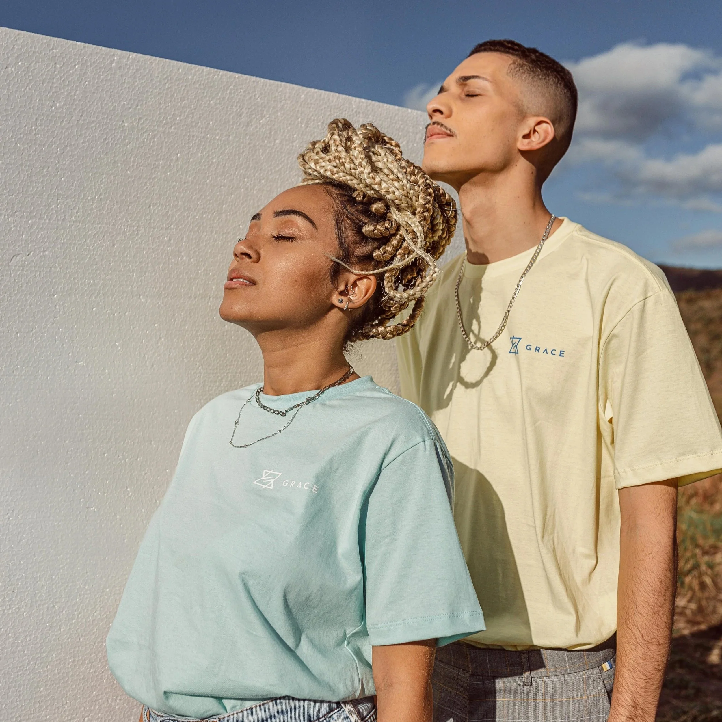 A woman with blonde braided hair and a man with short brown hair standing outdoors, both with eyes closed, facing the sun, wearing pastel-colored T-shirts with the word 'GRACE' printed on them.