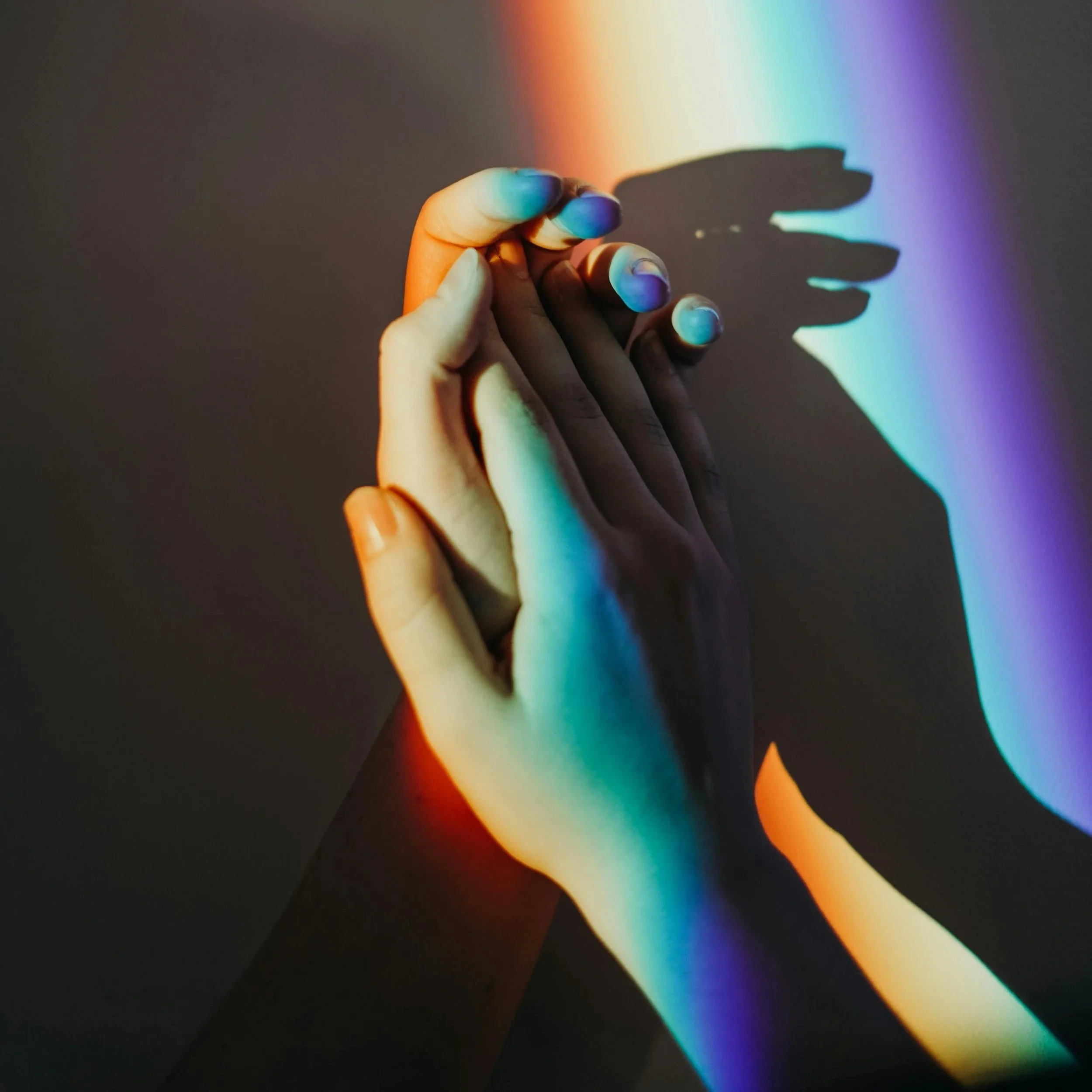 A hand with fingers intertwined against a plain background, with colorful rainbow light projection creating vivid shades and a shadow of the hand on the wall.