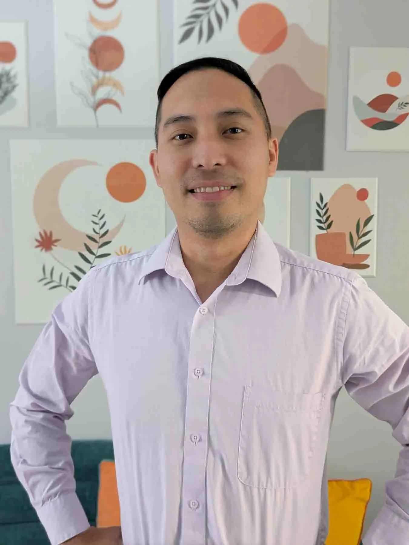 A man wearing a light pink button-up shirt smiling in front of a wall decorated with abstract art and graphics in earth tones.