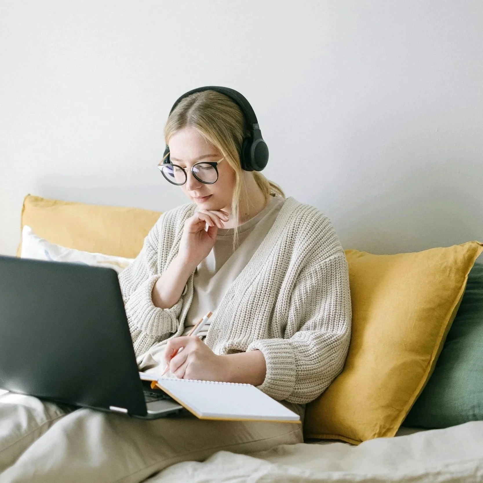 A woman with glasses and headphones sitting on a bed with yellow and green pillows, working on a laptop, holding a notebook and pen.
