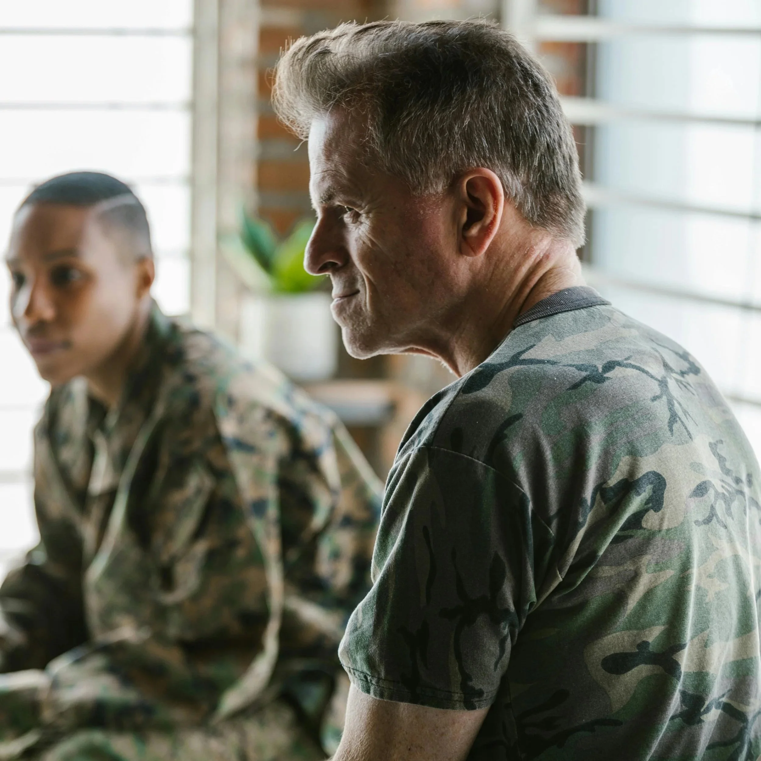 Two soldiers in camouflage uniforms sitting indoors near a window, engaged in a conversation.