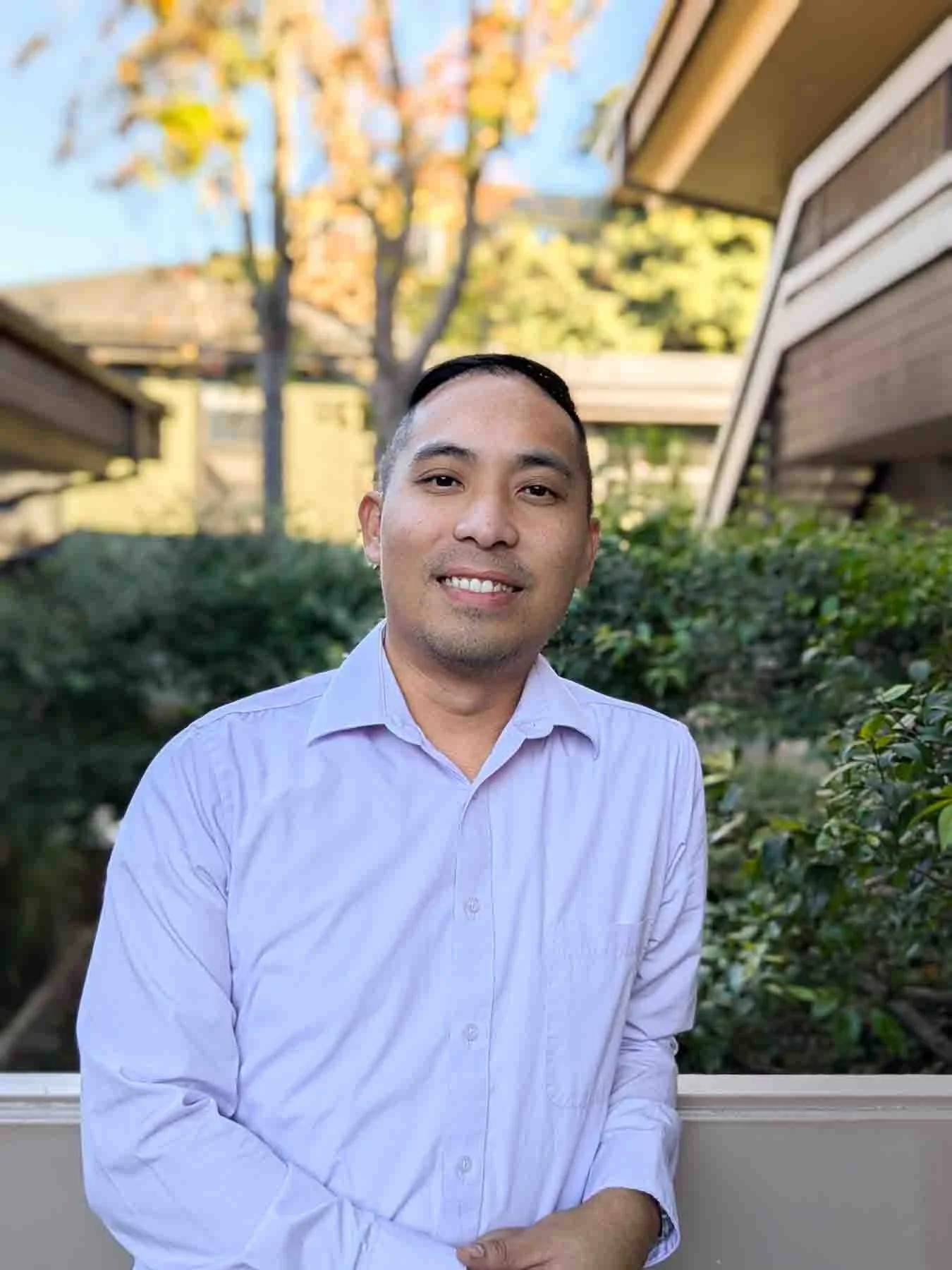 A man standing outdoors in front of green bushes, wearing a light purple button-up shirt, smiling, with residential buildings and a tree with yellow leaves in the background.