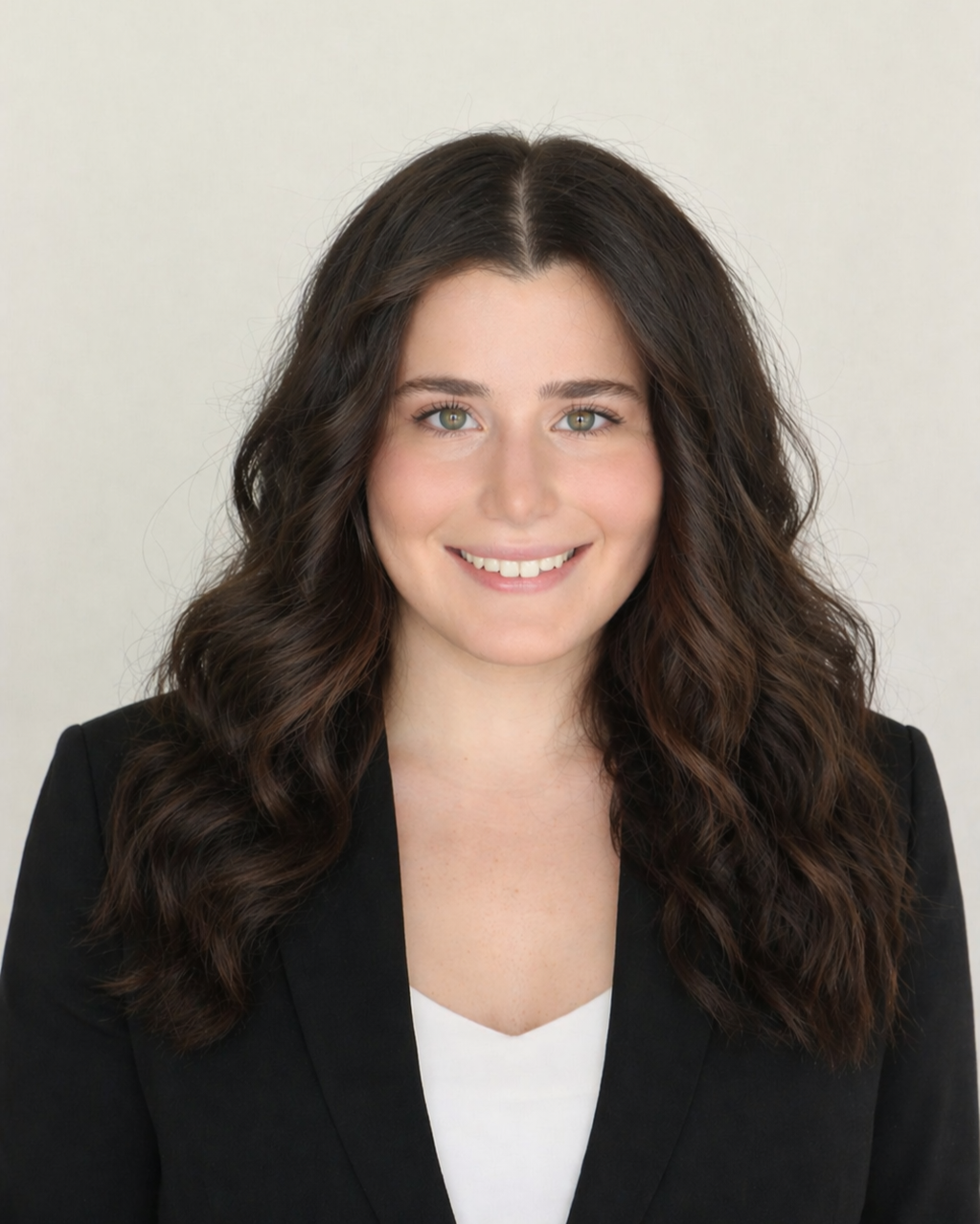Portrait of a young woman with long wavy brown hair, green eyes, and a bright smile, wearing a black blazer over a white top, against a light neutral background.