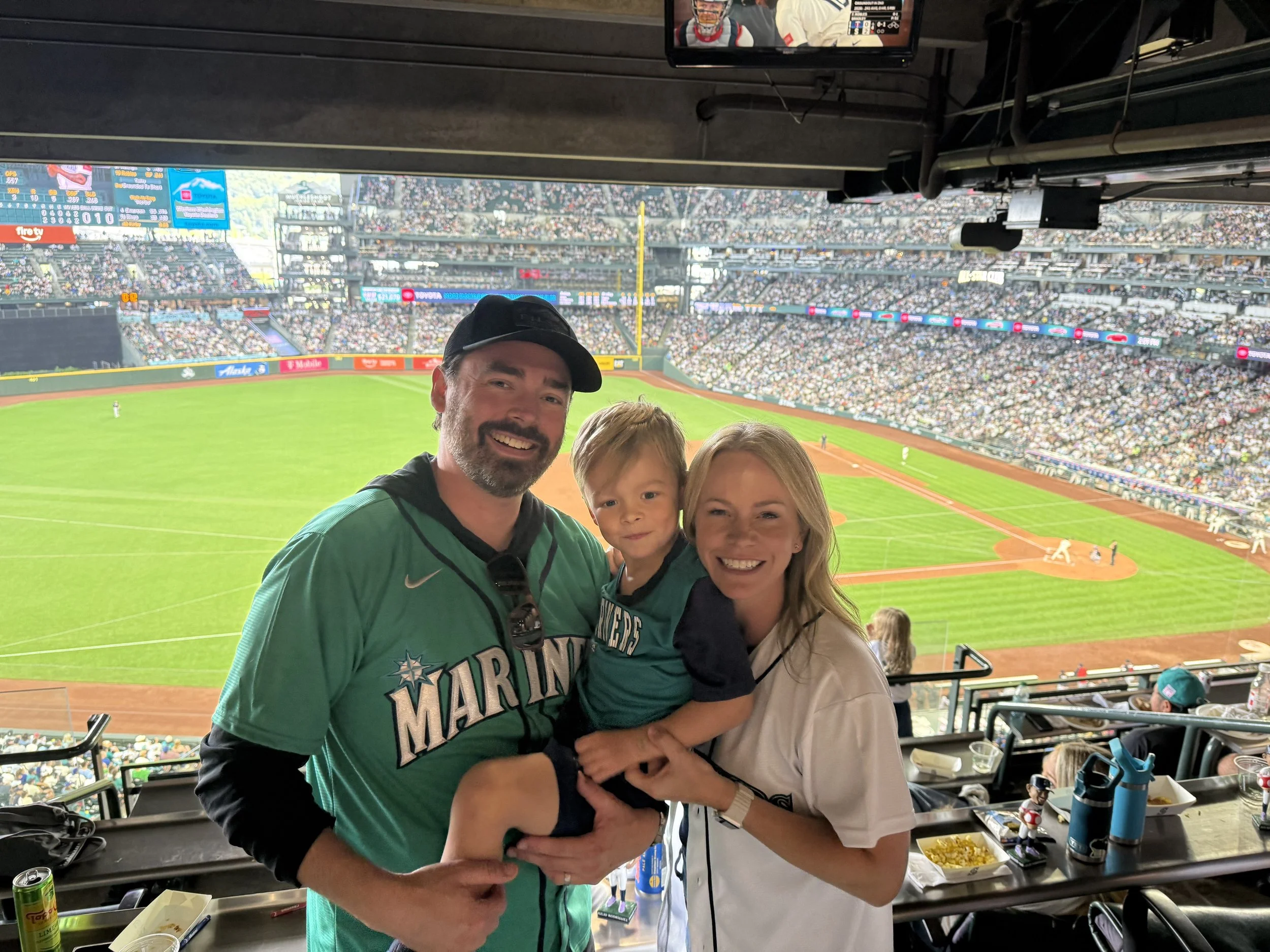 A family of four, including a man, woman, and two young children, pose for a photo in a baseball stadium's suite level, with the baseball field and stands filled with spectators visible in the background.