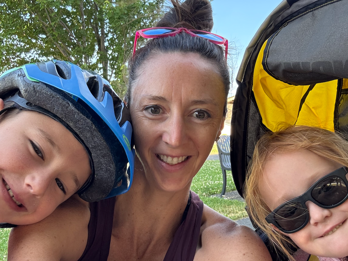 A woman with two children outdoors, one child wearing a bicycle helmet and the other wearing sunglasses, smiling for a selfie on a grassy area with trees and a park bench in the background.