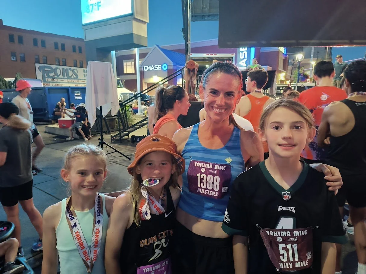 A woman and three young girls at a running event after finishing a race, smiling and posing for a photo, with medals around their necks, in an outdoor urban setting during evening.