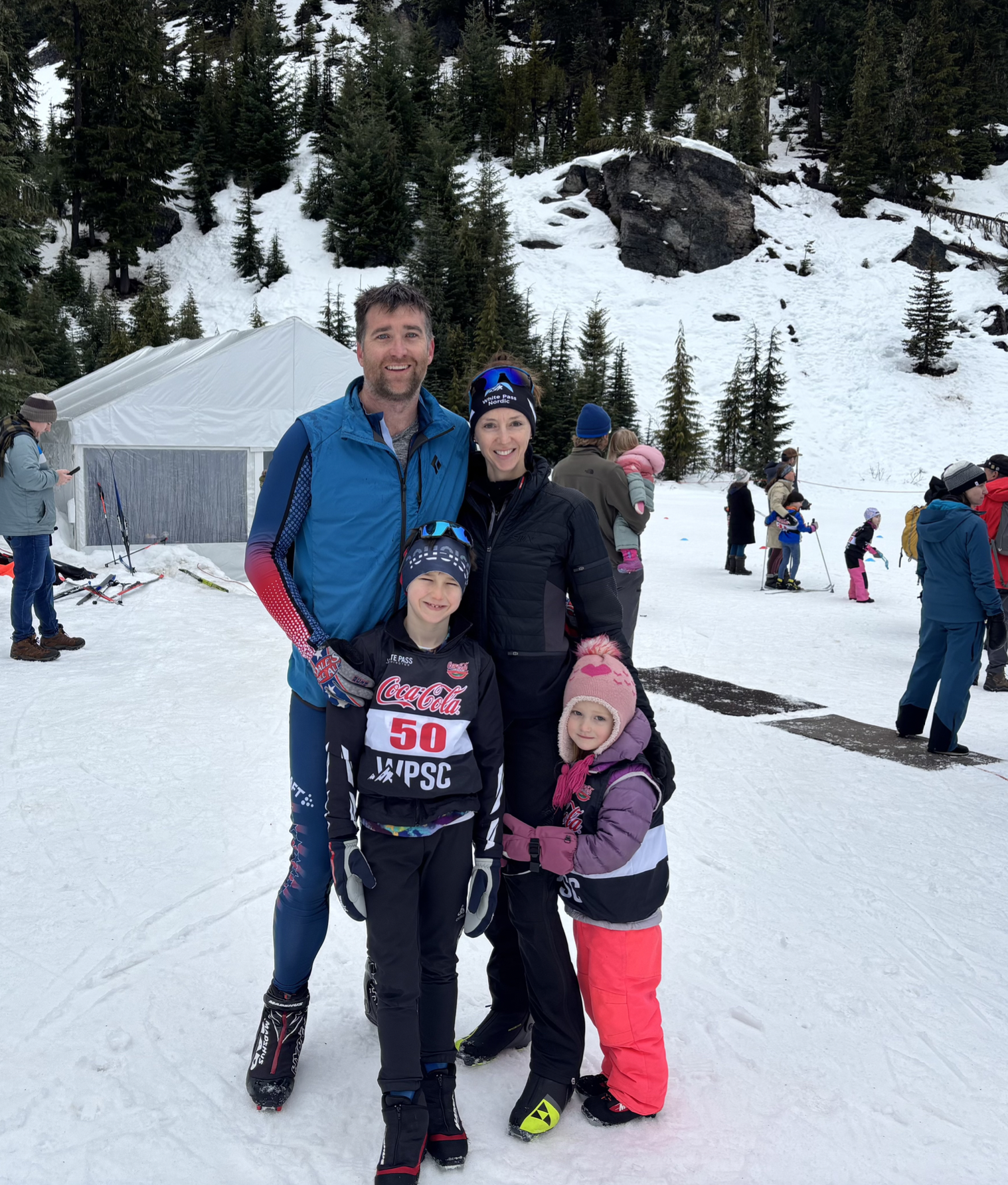 A family of four at a snowy mountain location, dressed in winter clothing with ski gear, standing and smiling for the photo. In the background, other people and children are seen enjoying winter activities, with a forested mountain behind them.