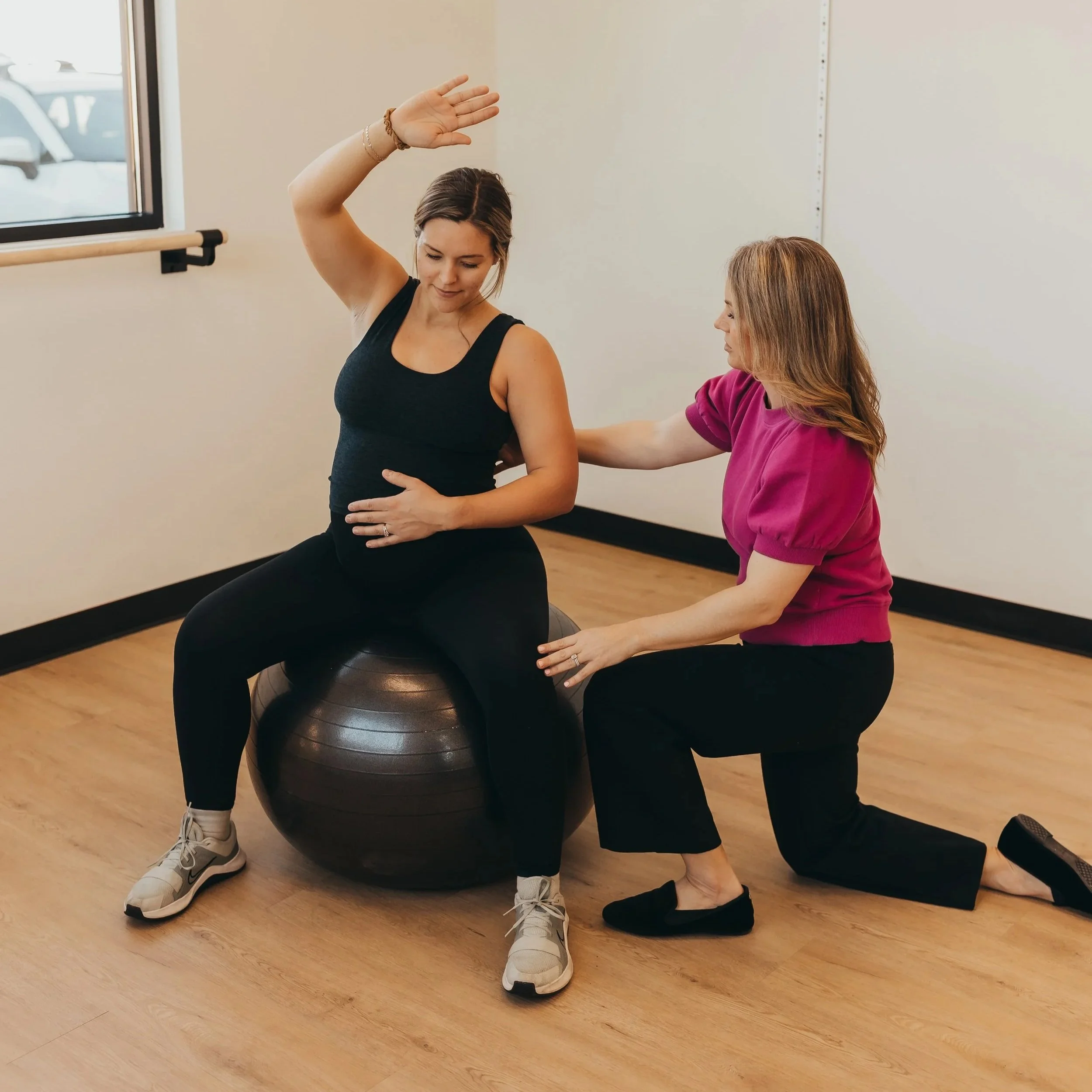 A woman in a purple shirt is assisting a woman in a black tank top with exercise on an exercise ball. The woman in black is raising her right arm. They are in a room with wood flooring and a ballet barre near a window.