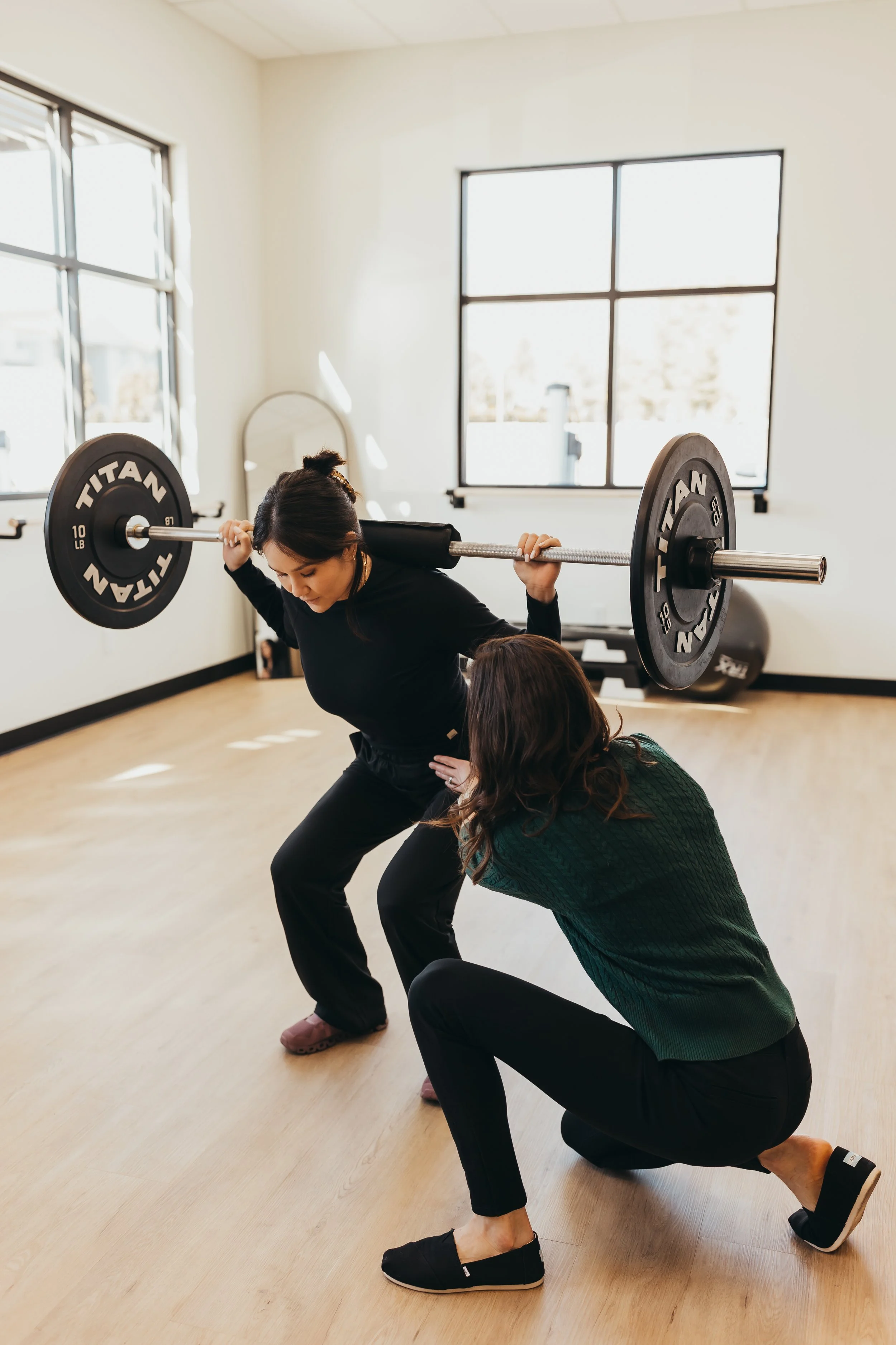 A woman is performing a squat exercise with a barbell on her shoulders while a trainer supports her in a gym with large windows and wooden flooring.