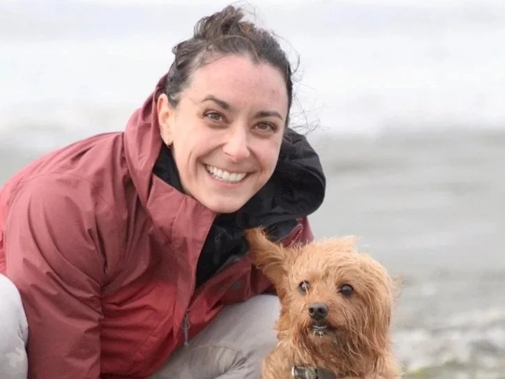 Woman smiling with a small brown dog near a body of water.