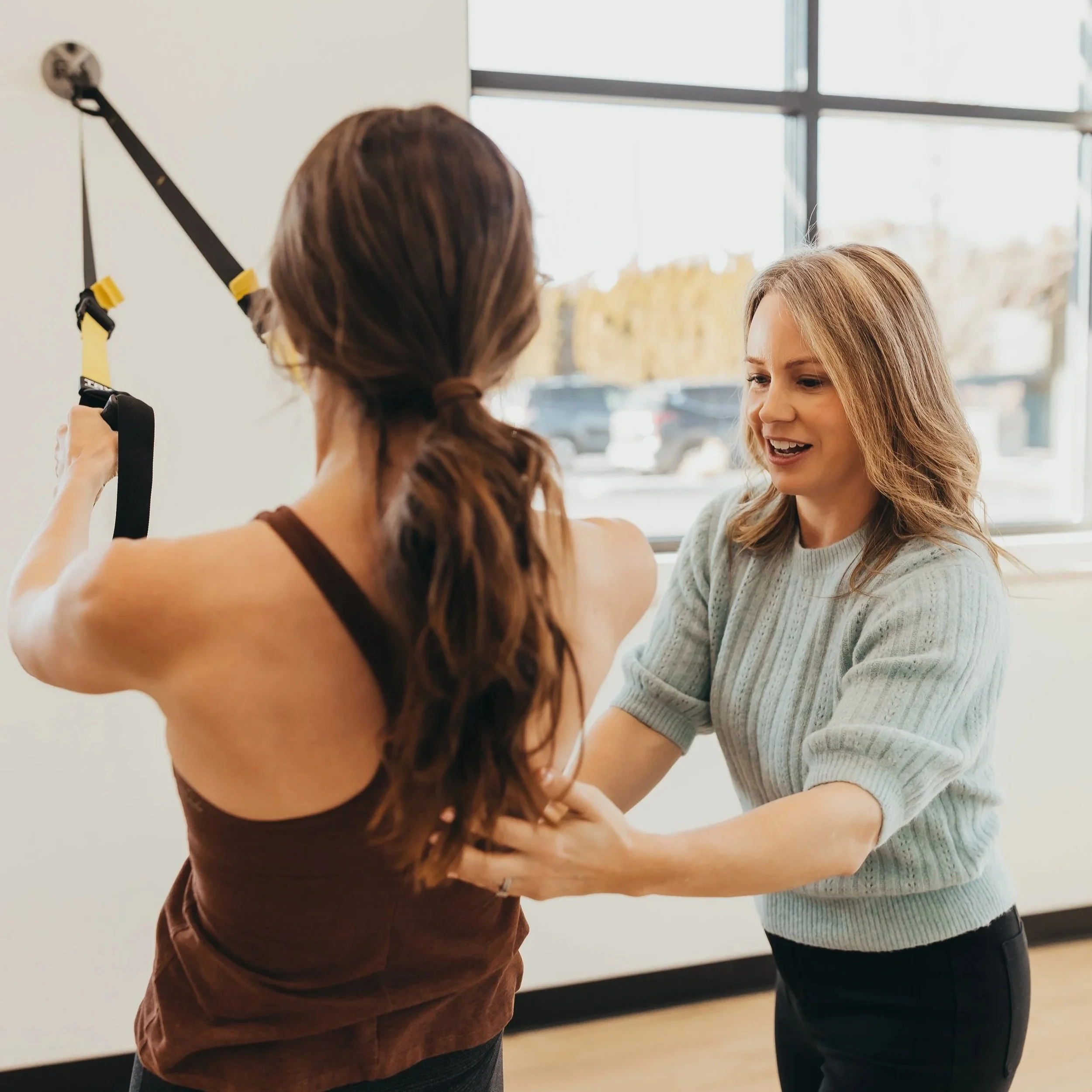 Two people are in an indoor setting, with one person performing a fitness or physical therapy exercise using a strap or resistance band while the other person observes or guides. The focus is on their interaction and the activity they are engaged in.