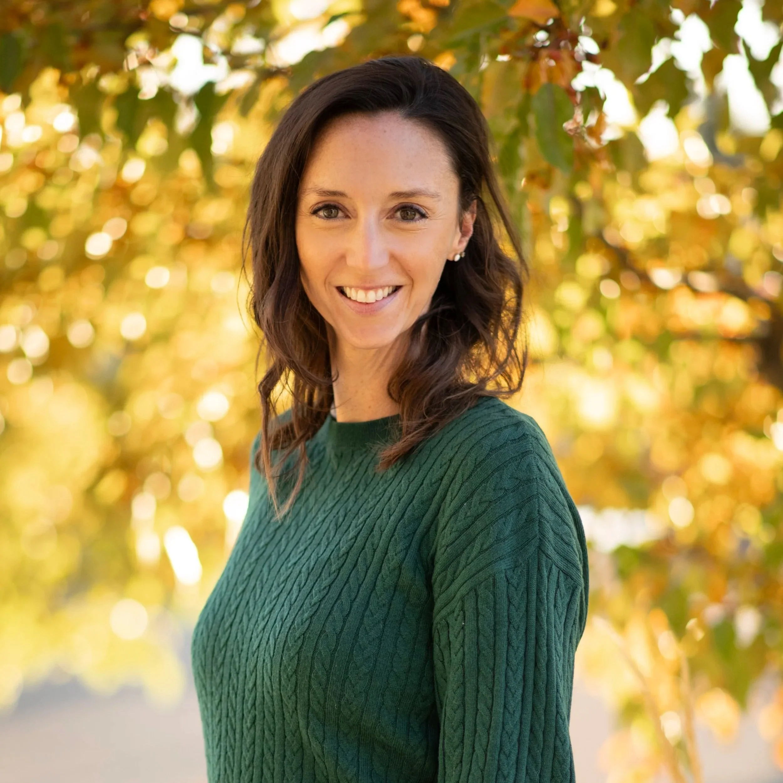 A smiling woman with dark wavy hair wearing pearl earrings and a green textured sweater, outdoors during autumn with blurred yellow and brown leaves in the background.