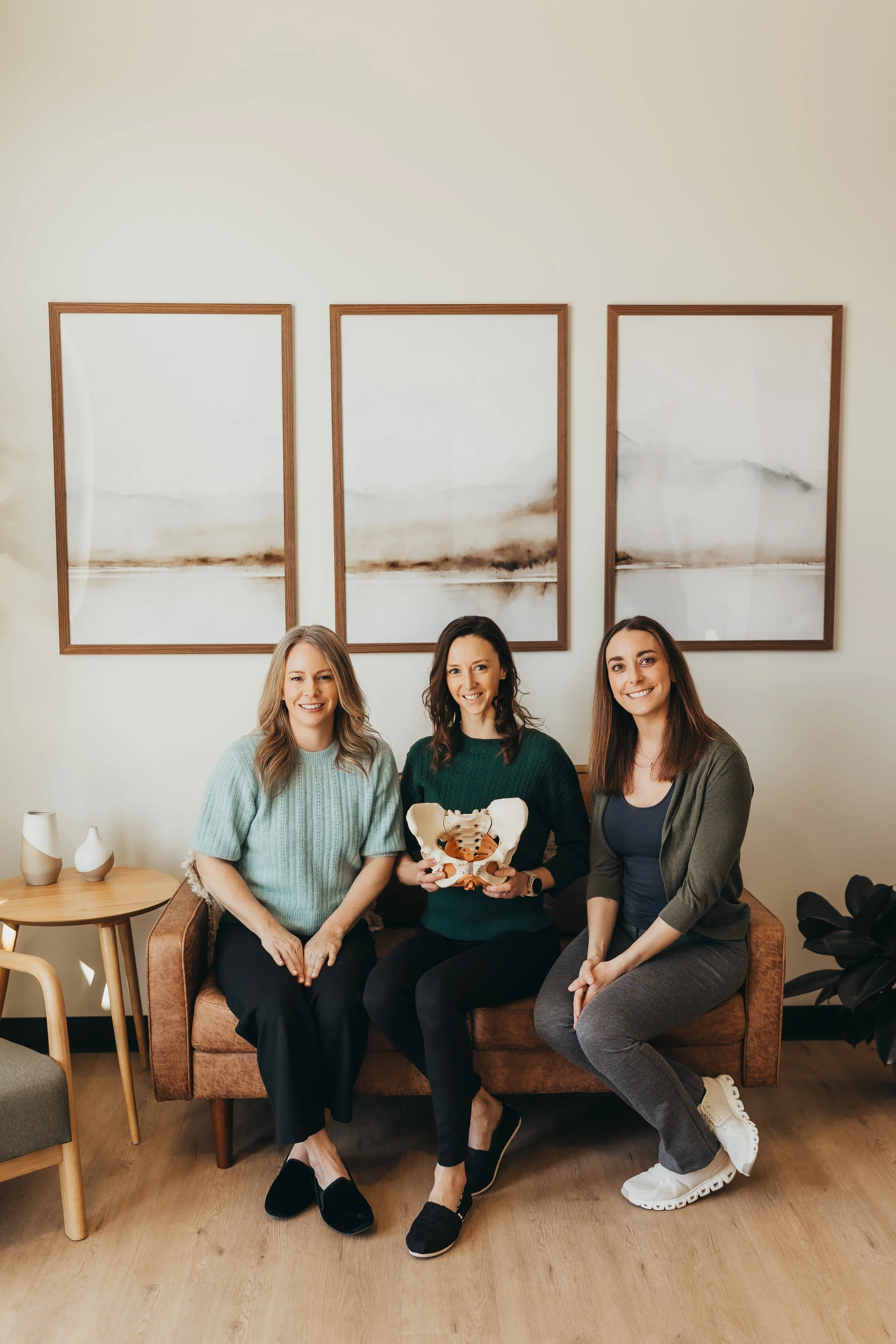 Three women sitting on a brown couch with a wooden table and vases to their left, one woman in the middle holding a pelvic model. Behind them, three abstract artworks hang on a white wall, and a black plant is visible on the far right.