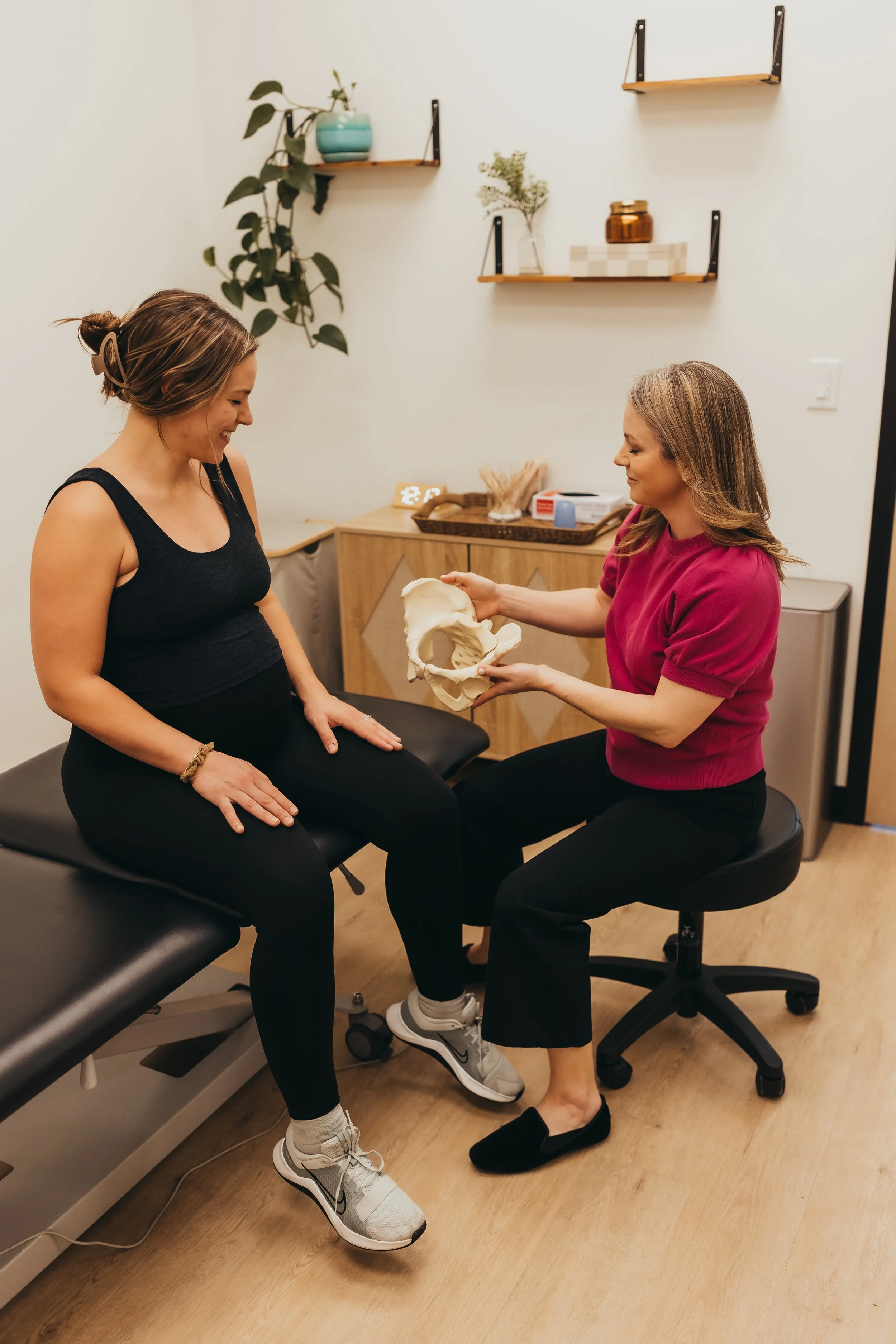 A pregnant woman having a pelvic exam with a healthcare professional in a medical office, the healthcare professional holding a pelvic model while sitting on a rolling stool.