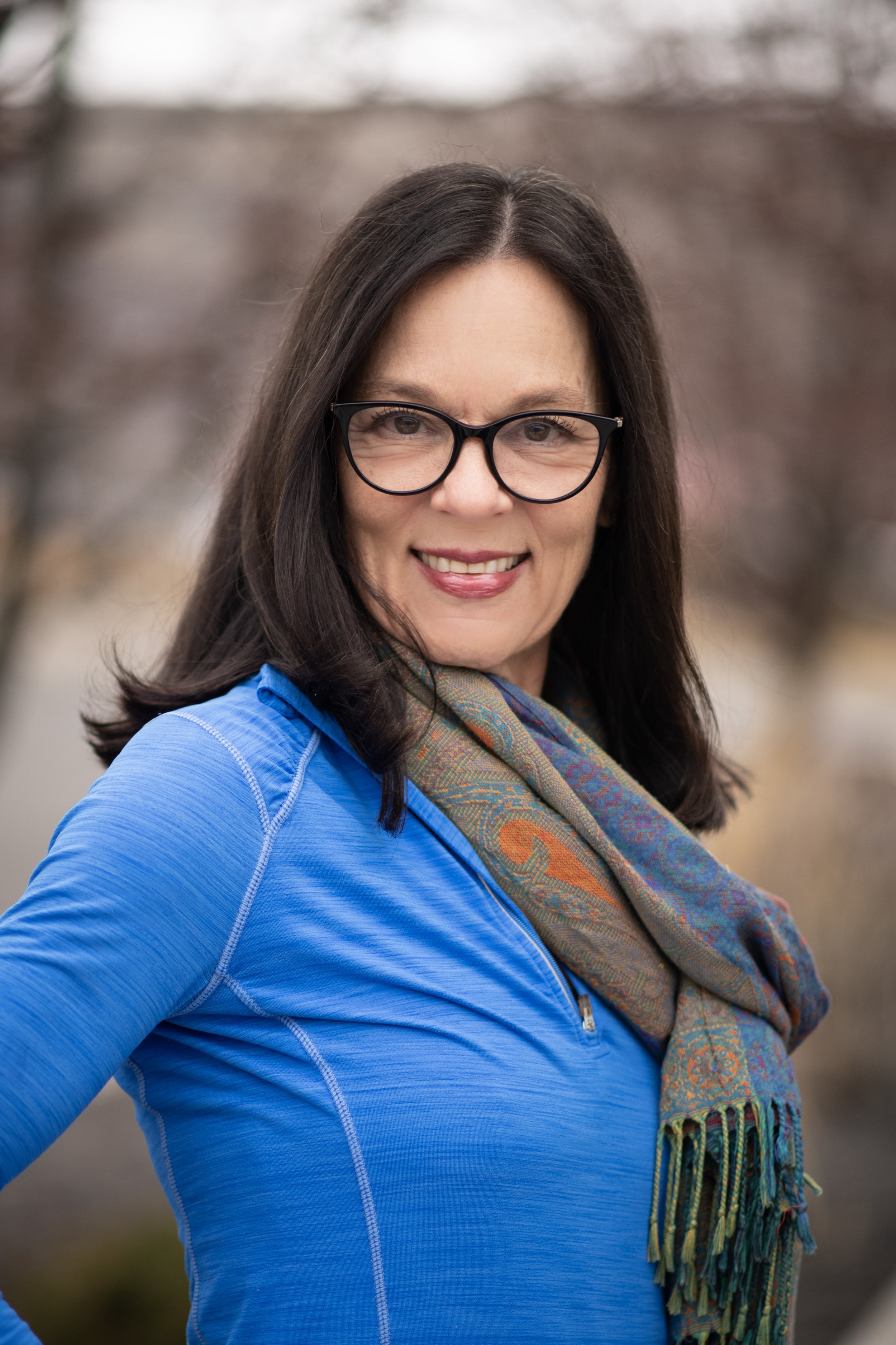A woman with long dark hair, glasses, and a bright blue jacket standing outdoors with a blurred natural background, smiling at the camera.