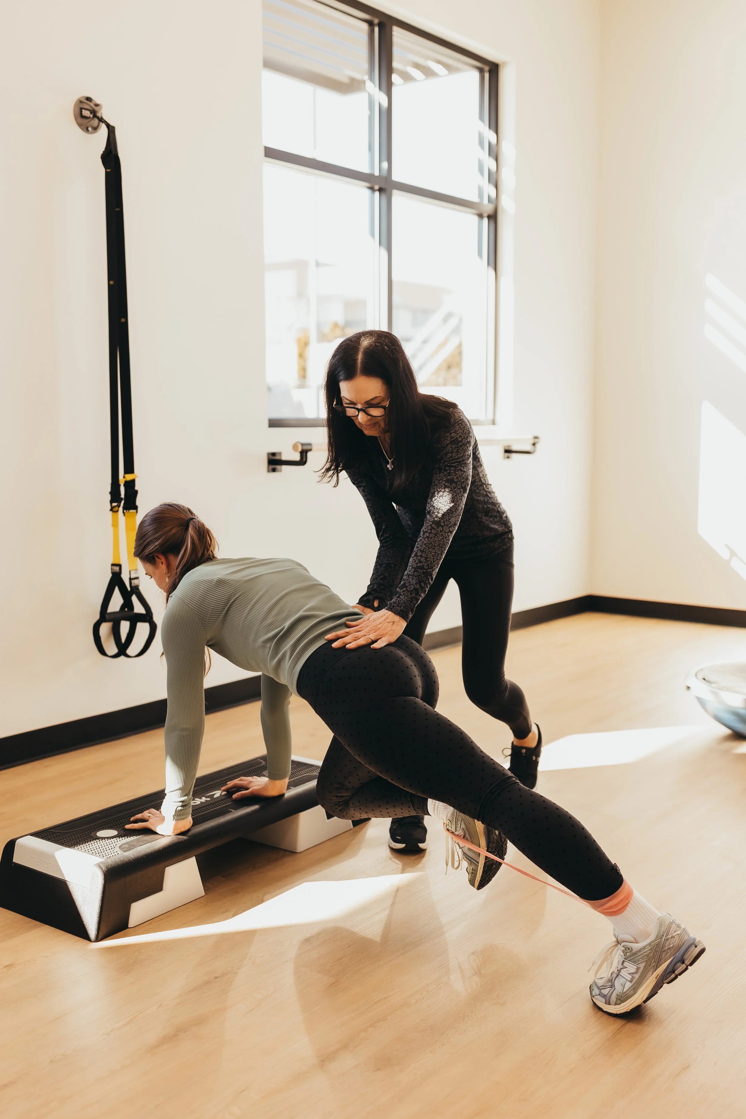 A woman is performing a plank exercise on a workout step, while a trainer supports her with one hand on her back. The setting is a bright fitness room with a large window.