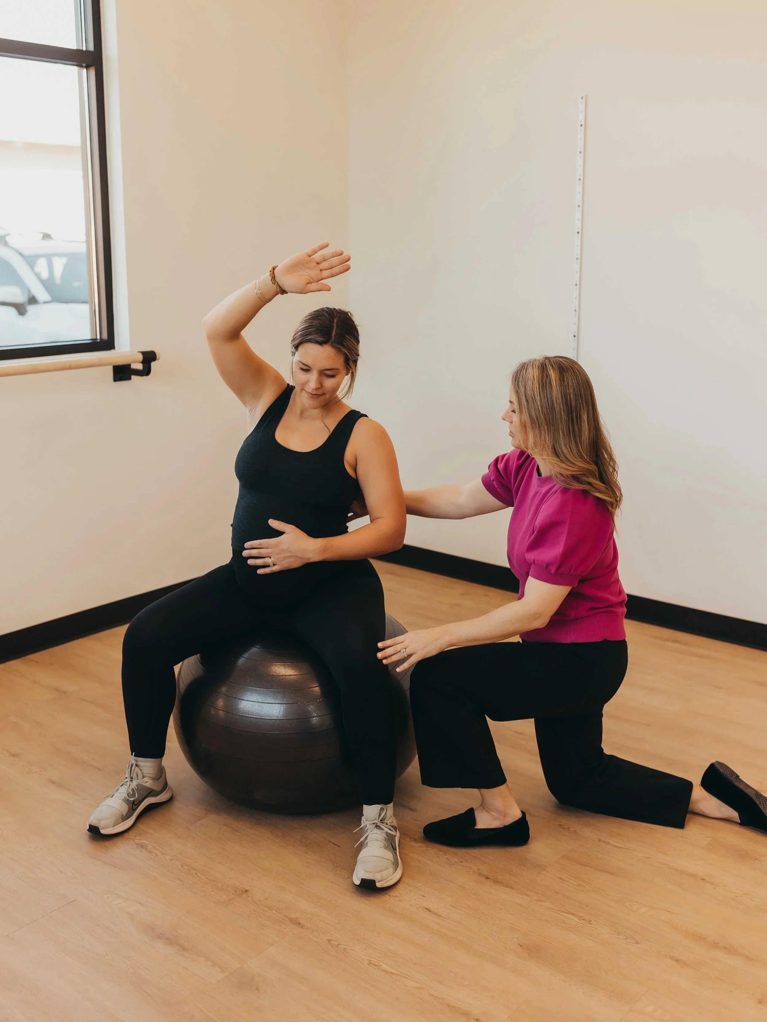 A pregnant woman sitting on an exercise ball with her left arm raised, while a trainer kneels beside her supporting her by her right arm and hand. The scene is in a fitness or physical therapy studio.