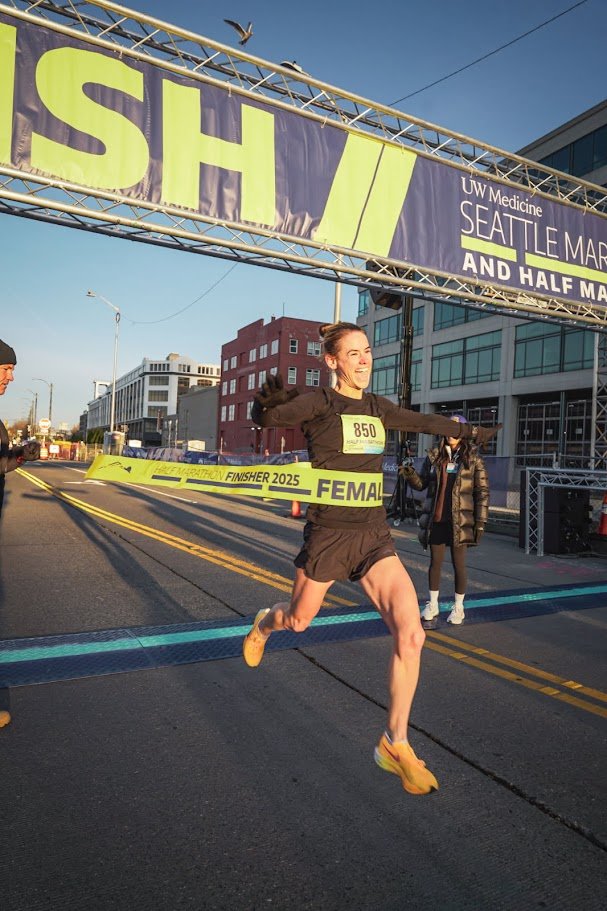 Female marathon runner crossing the finish line at a race event in an urban area, with a finish line banner overhead and city buildings in the background.