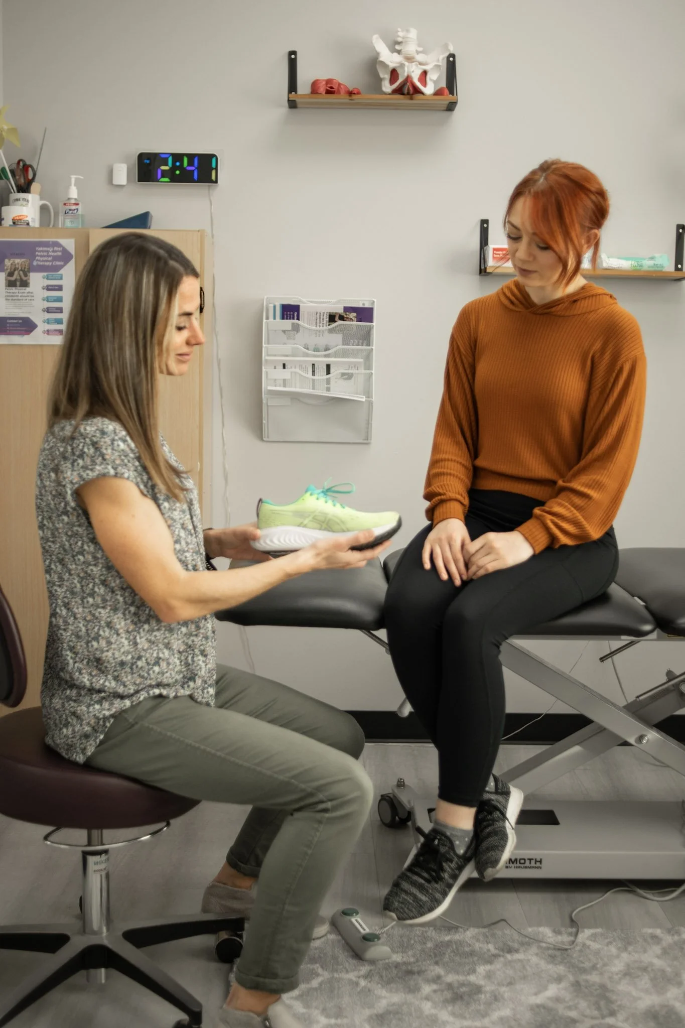 A woman sitting on an examination table in a medical office while another woman shows her a running shoe. The woman on the table is wearing a brown hoodie and black pants, the other woman is seated on a stool, wearing a patterned blouse and green pants. A scale is on the floor, and a wall clock shows 2:41. Medical supplies and anatomical models are visible in the background.