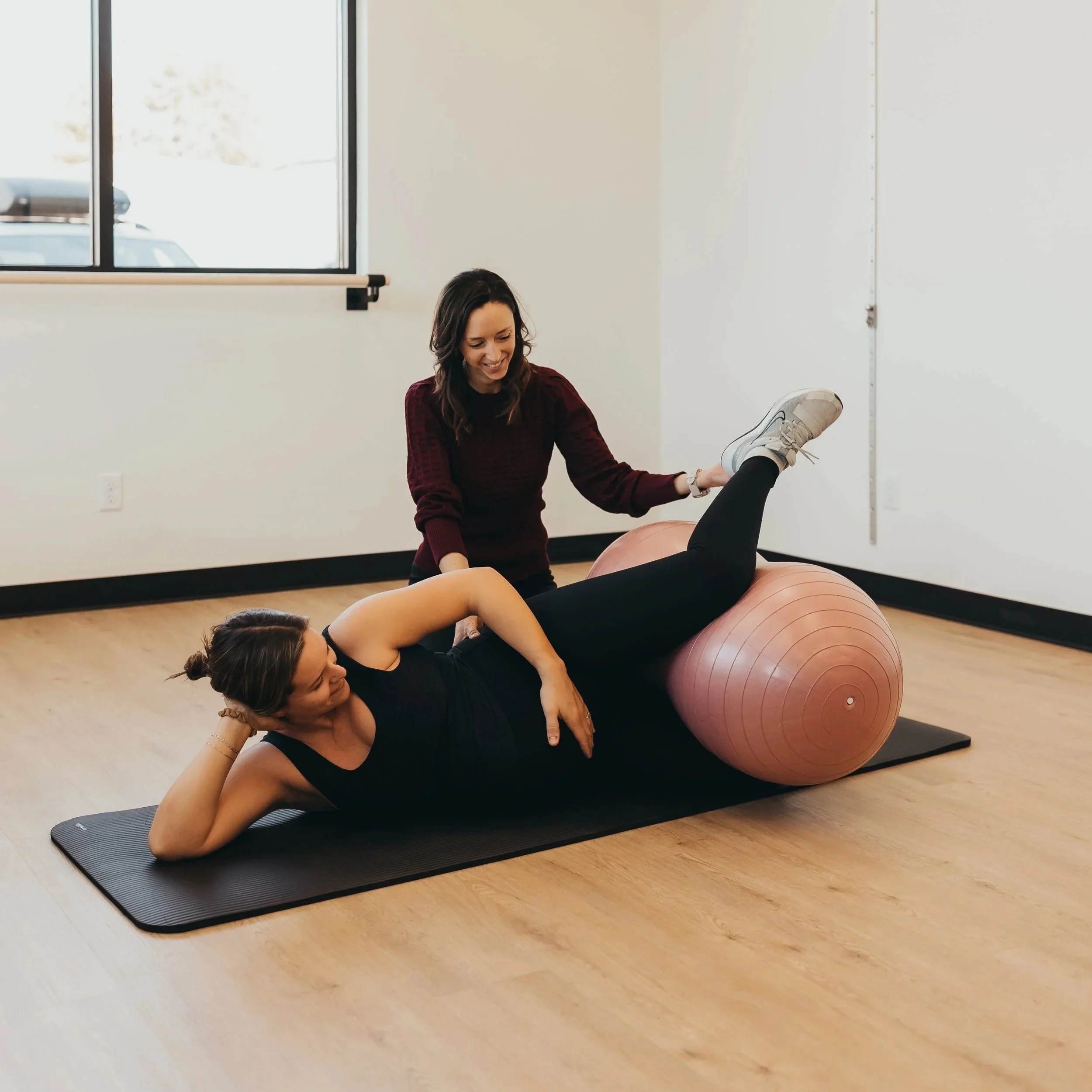 A woman in black leggings and a black tank top lying on a black exercise mat with a pink exercise ball under her legs, while another woman in a maroon sweater supports her during a stretch or exercise session in a bright room with large windows.