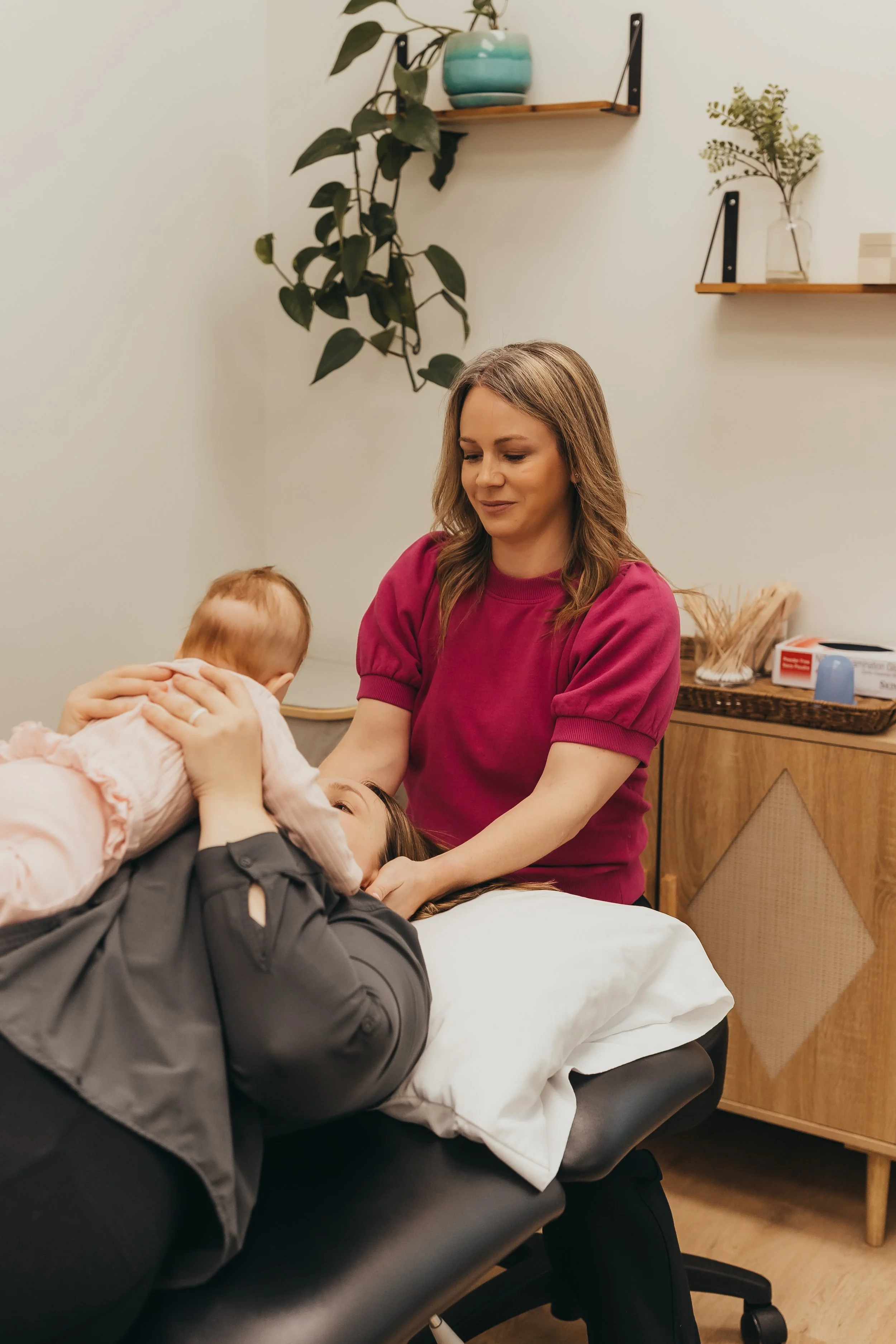 A woman lying on a treatment table holding a baby, with another woman, likely a healthcare provider, gently supporting the baby's head, in a room with wall-mounted shelves and potted plants.