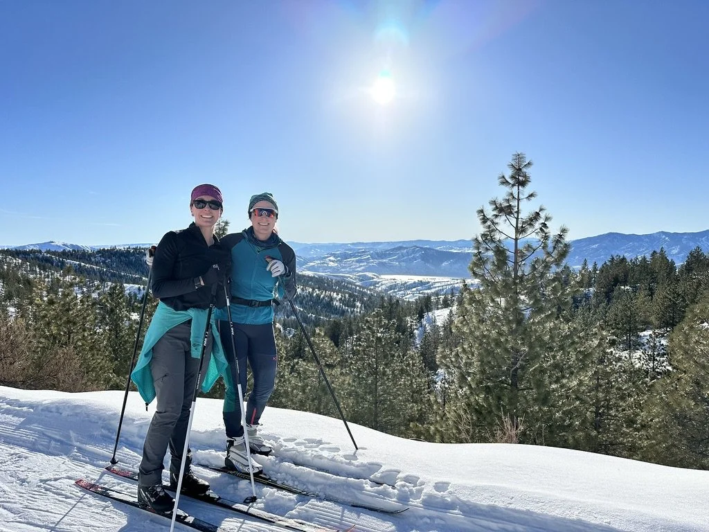 Two women in winter gear smiling and posing with ski poles on snowy mountain with trees, mountains, and clear blue sky in the background.