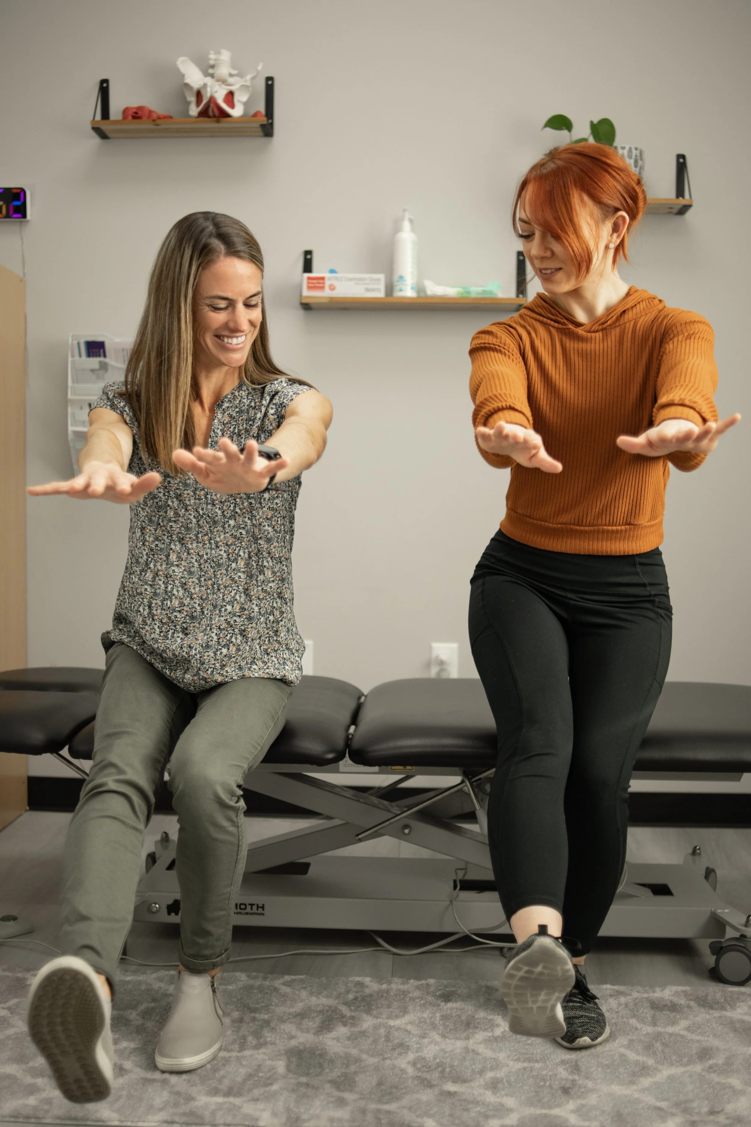 Two women performing leg and arm stretches in a medical or physical therapy office, with a medical examination table and shelves with medical supplies in the background.