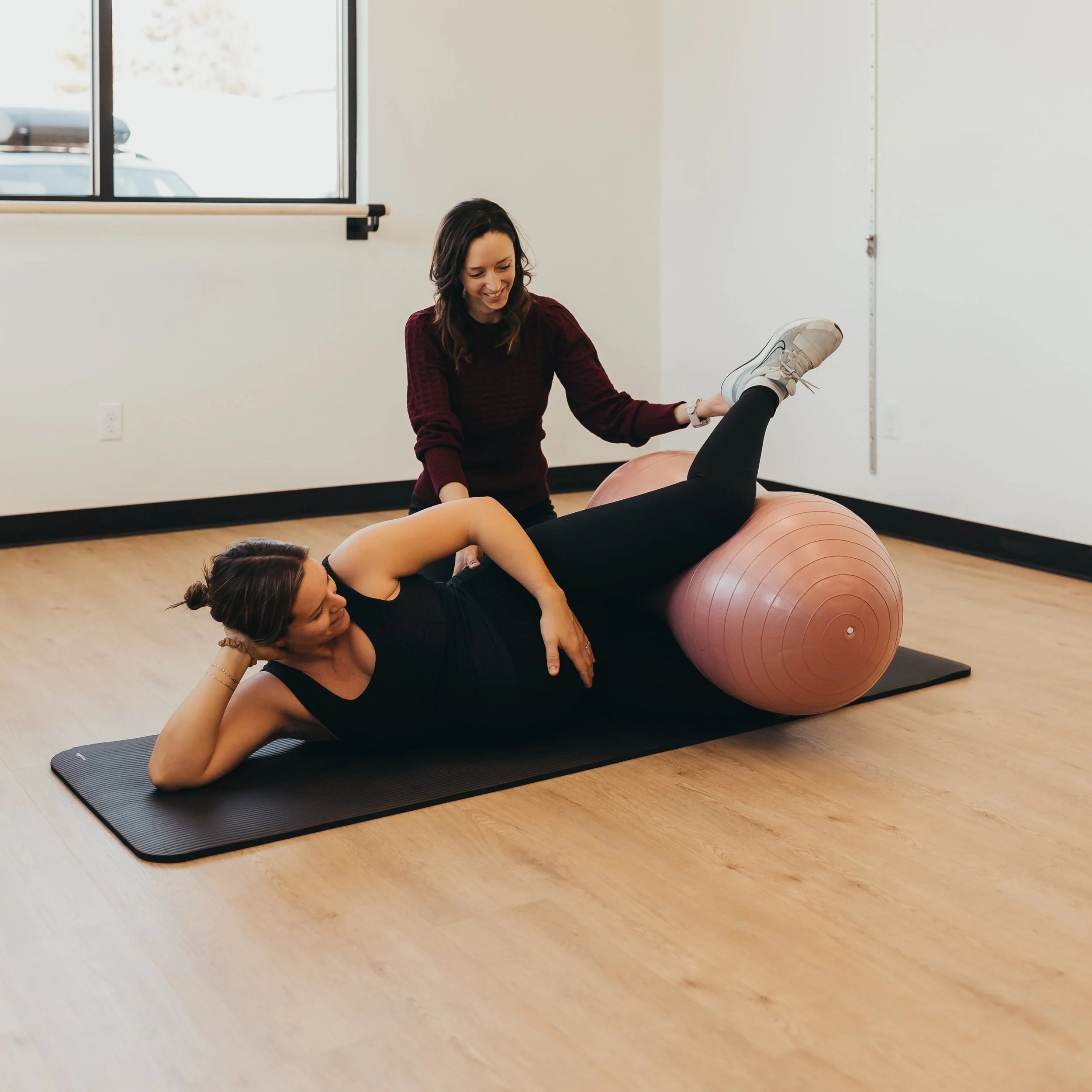 A woman assisting another woman during a physical therapy session on an exercise ball; exercise mat and window in background.