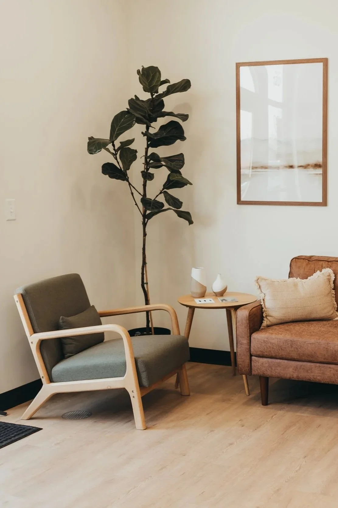 Living room corner with a tall fiddle leaf fig plant, a beige armchair with a gray pillow, a rust-colored sofa with a beige pillow, a round wooden side table with two ceramic vases, and a framed artwork on the light-colored wall.