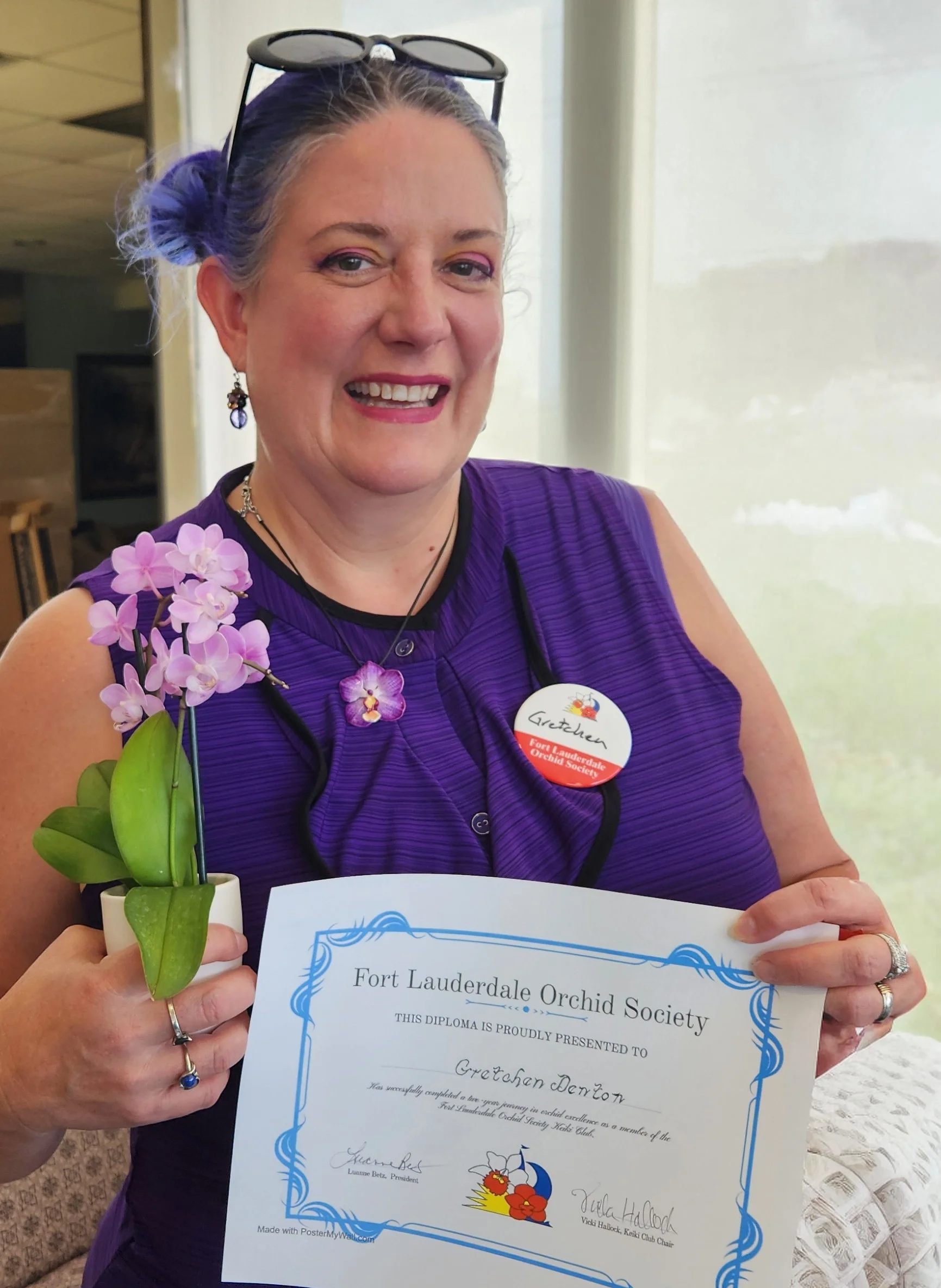 A woman with purple hair styled in buns, wearing a purple sleeveless top, holding a diploma from Fort Lauderdale Orchid Society, and a small orchid plant with pink flowers and green leaves. She is smiling and has glasses on her head, earrings, rings, and a necklace. She also has a badge that says 'Gretchen'.
