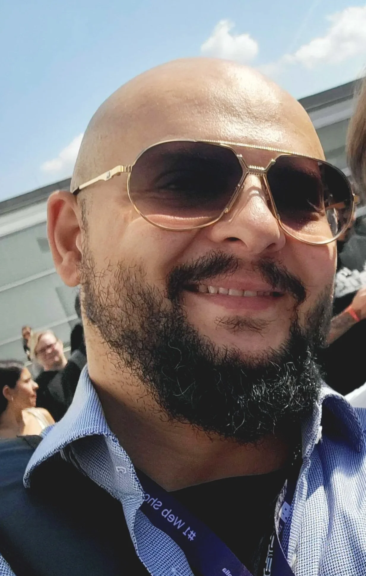 Close-up of a smiling man with a beard wearing Cazal sunglasses, a blue collared shirt, and a lanyard around his neck, outside during daytime with a building and a few people in the background.