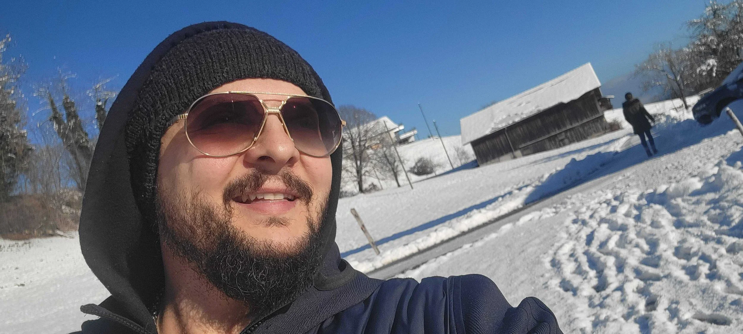 Close-up of a bearded man wearing sunglasses, a black beanie, and a jacket, smiling while taking a selfie in a snowy landscape with a building, trees, and another person walking in the background.