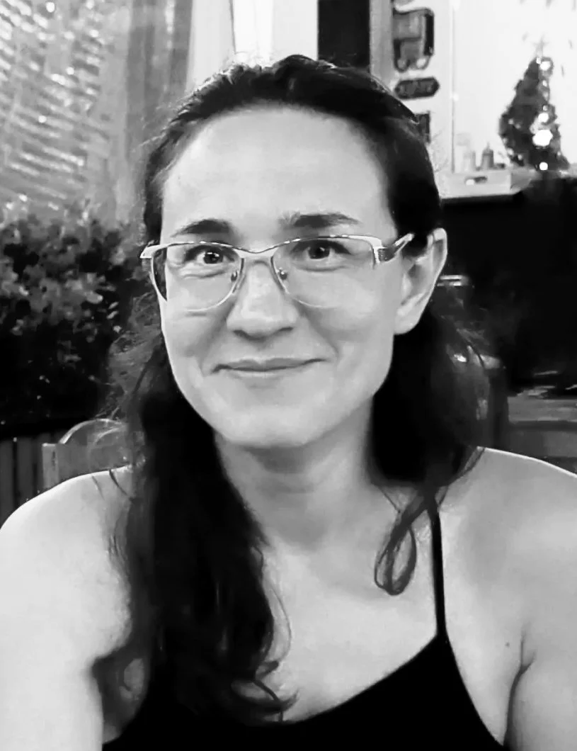 A black and white photo of a smiling woman with glasses, long dark hair, wearing a sleeveless top, sitting indoors with decorative items and a small Christmas tree in the background.
