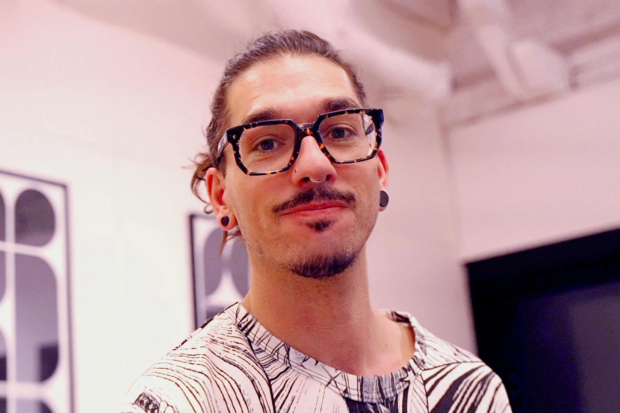 A young man with long brown hair tied back, wearing large tortoiseshell glasses, a nose ring, ear gauges, and a patterned shirt, standing indoors with a neutral expression.